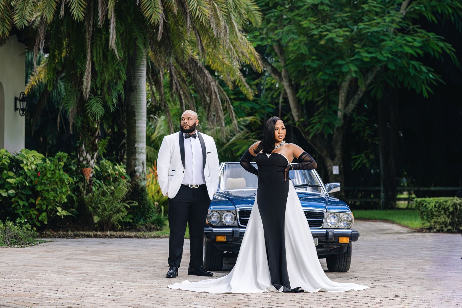 Elegant couple in black and white formal attire posing in front of a vintage blue car surrounded by lush tropical greenery.