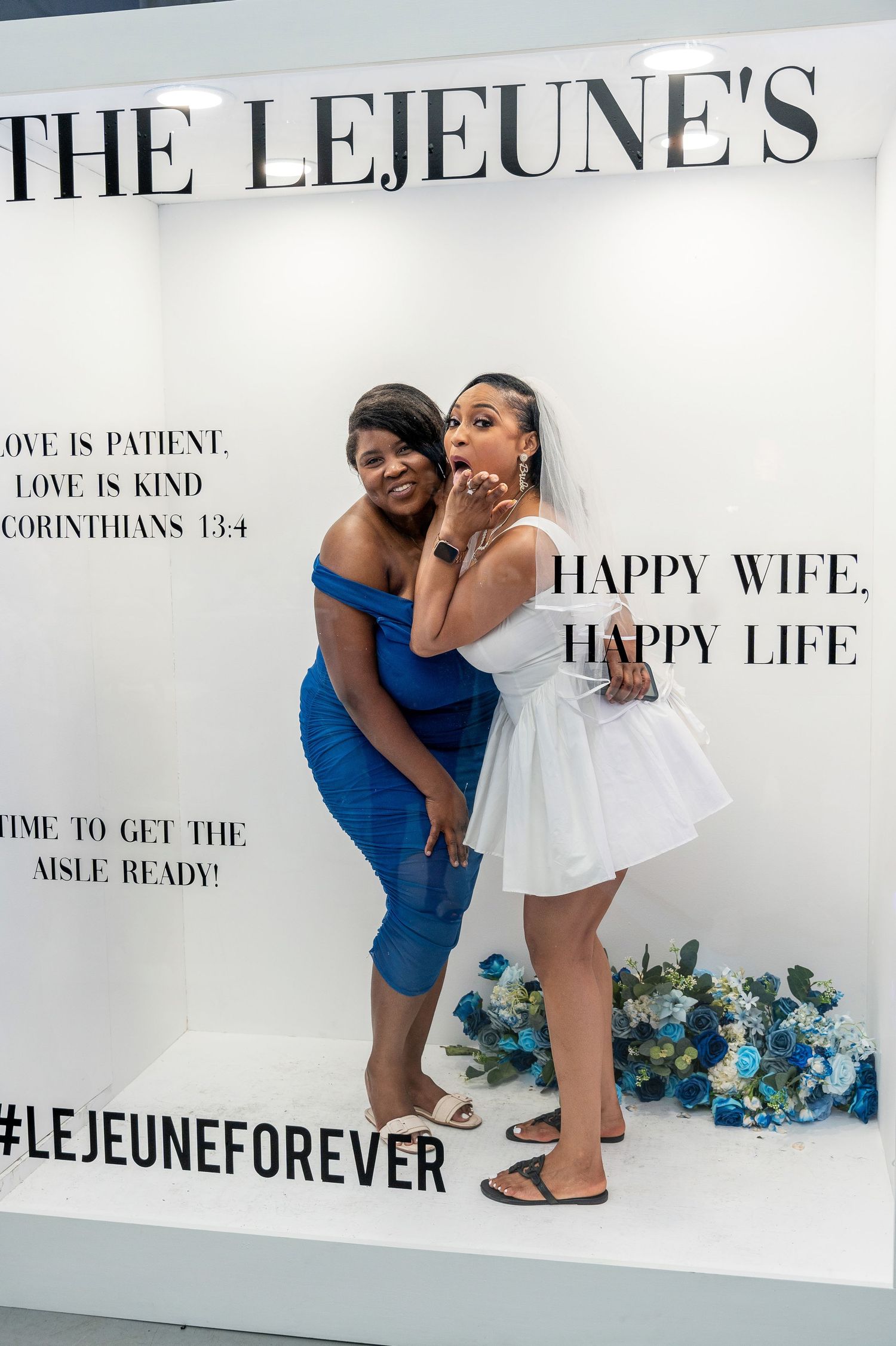 Bride in white mini dress and veil poses with woman in blue dress inside The Lejeune's photo booth.