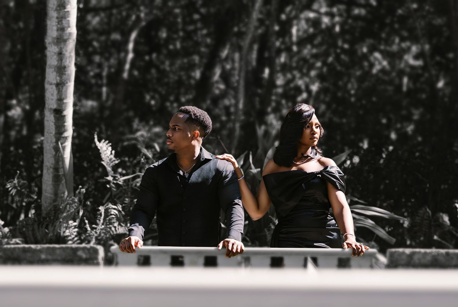 Black and white photo of couple leaning on bridge railing in outdoor setting.