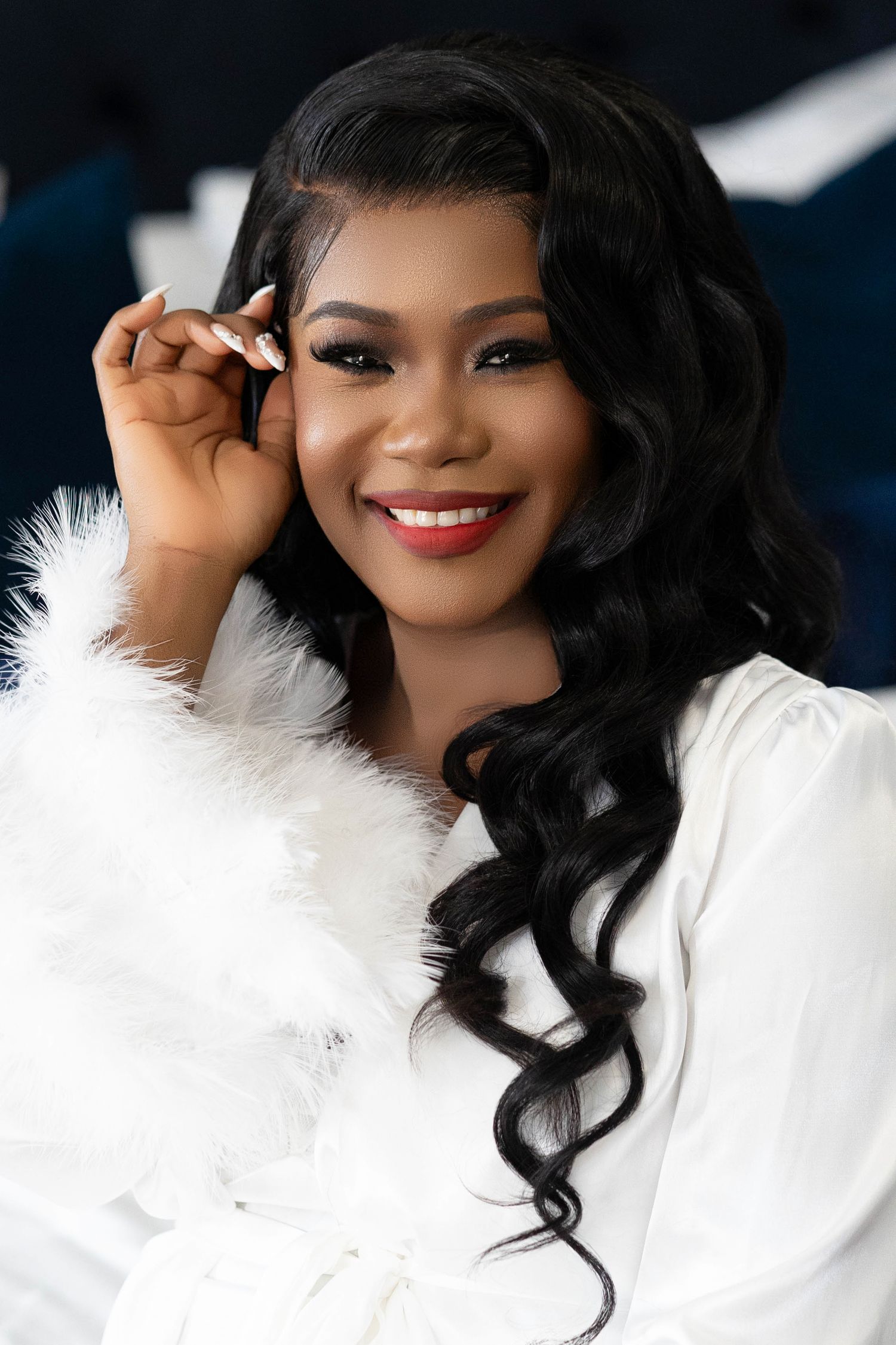 Smiling Black woman with long curly hair wearing a white feather robe and red lipstick poses elegantly.