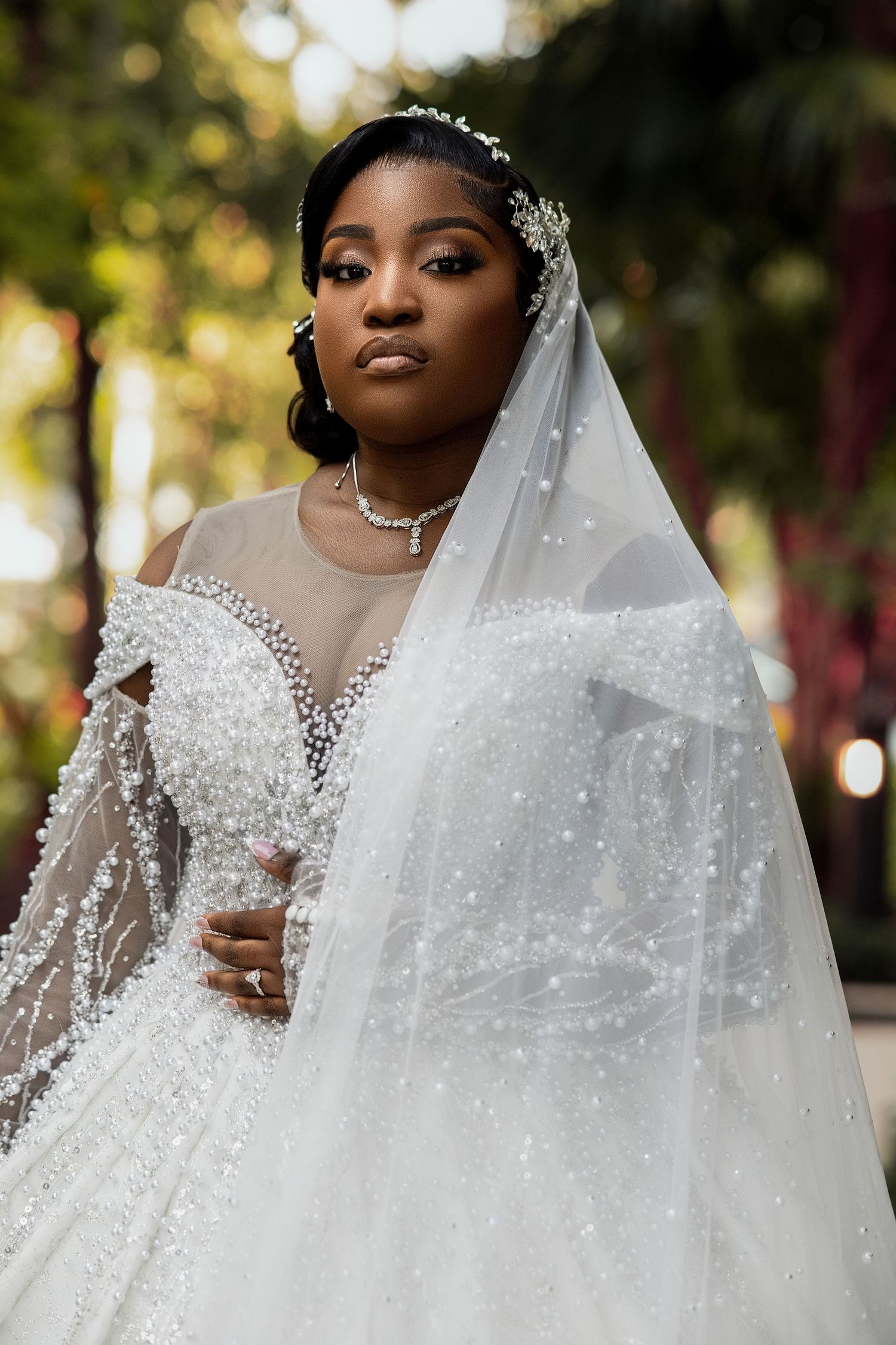 A Black bride in a pearl-embellished ball gown and veil poses outdoors, wearing a diamond necklace and floral hair piece.