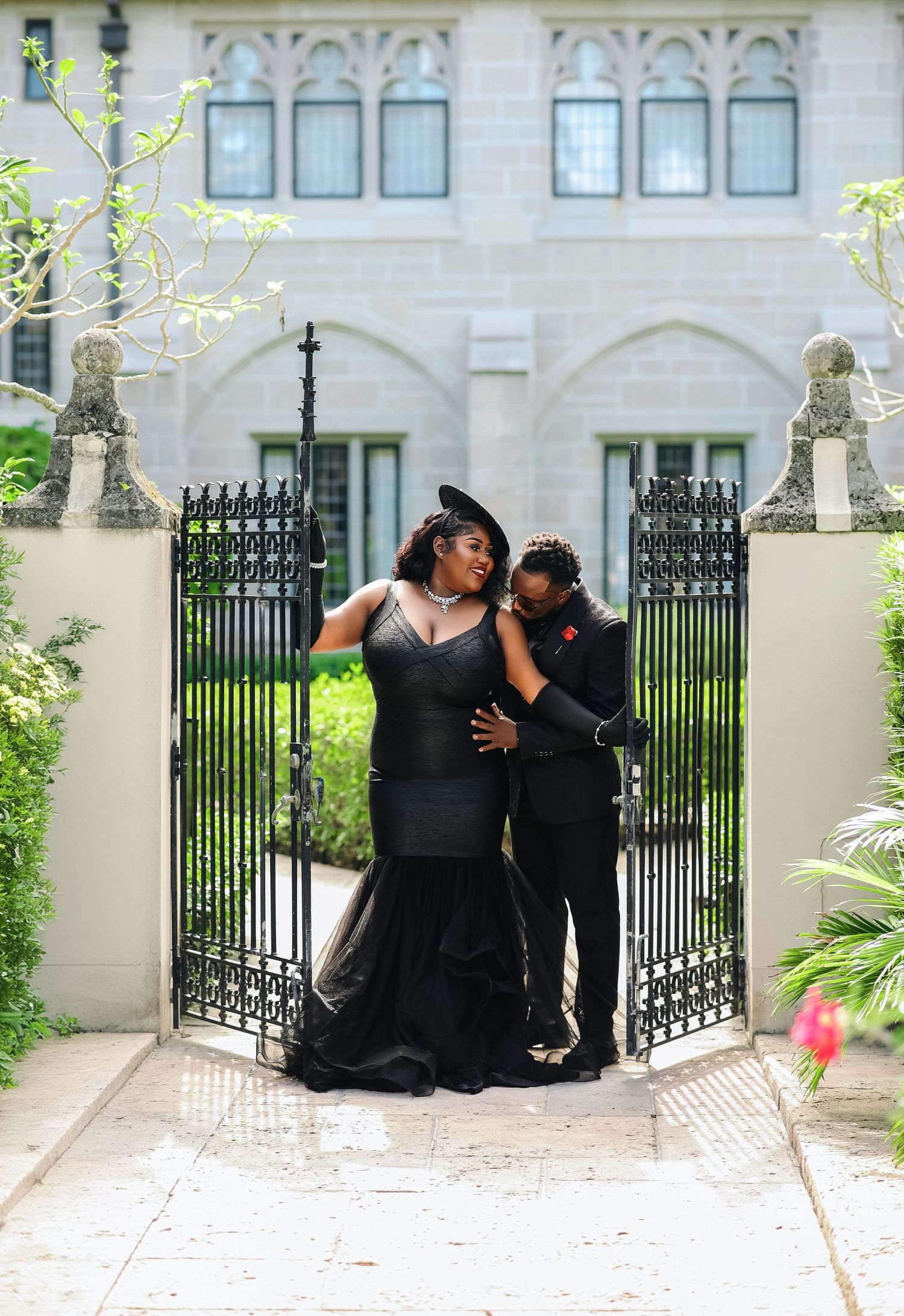 A couple in black formal wear share a romantic moment framed by ornate iron gates of a historic mansion.
