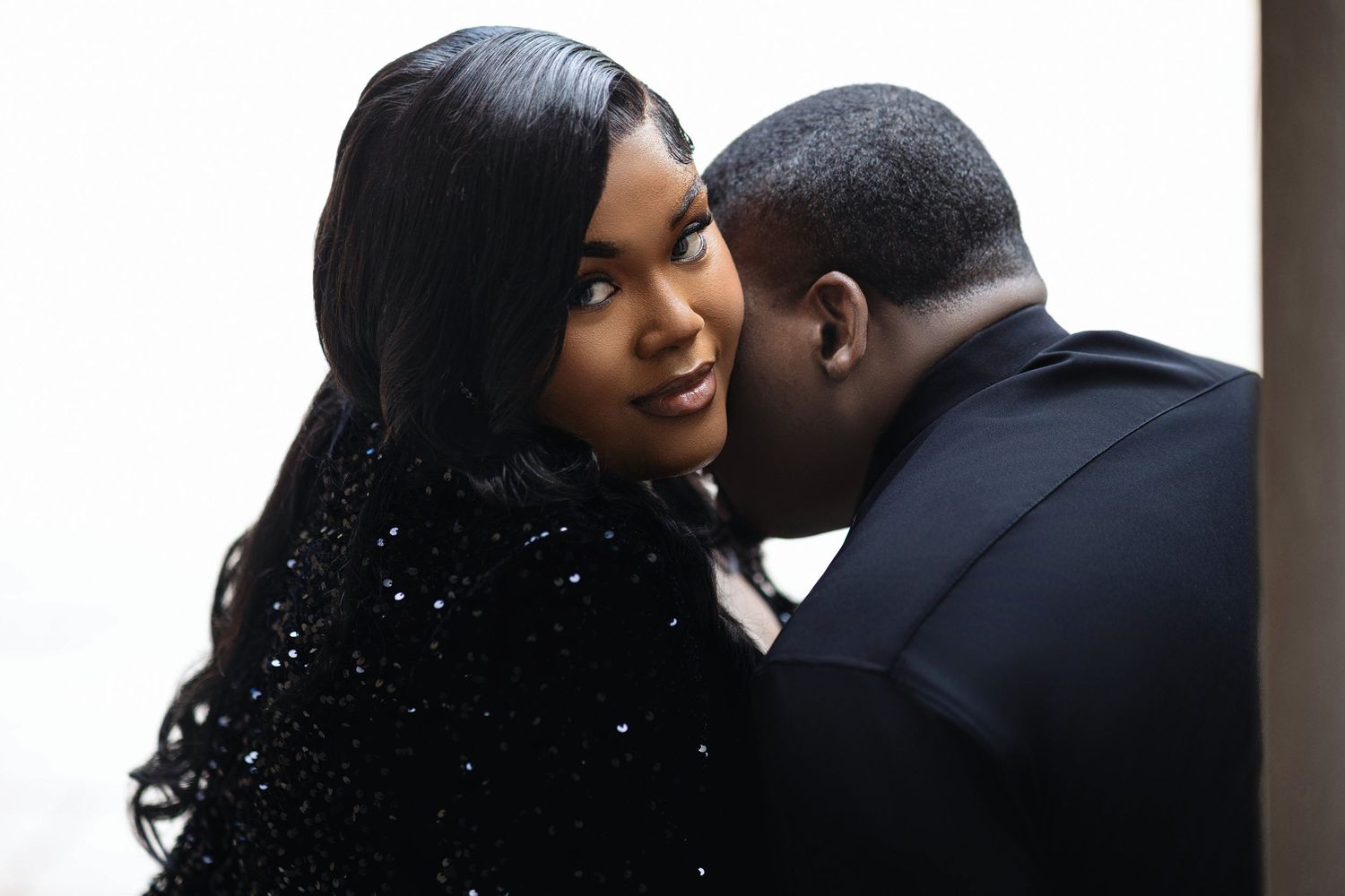 Romantic couple sharing a tender moment with man whispering to smiling woman in black sequin dress.