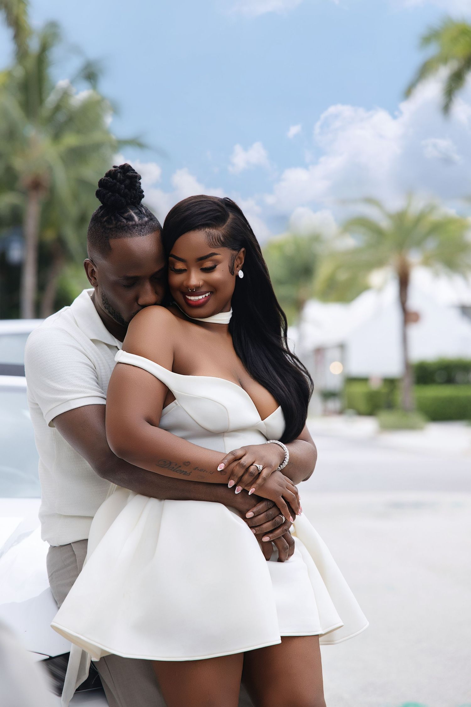 A Black couple shares a warm embrace near palm trees, with the woman wearing a white mini dress.