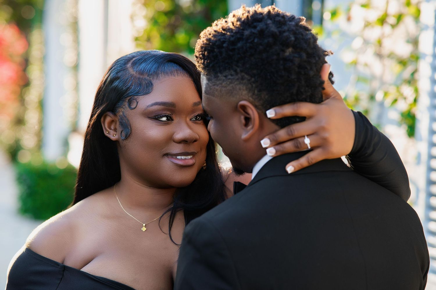 A couple in black formal wear share a tender moment, the woman smiling as she embraces her partner outdoors.