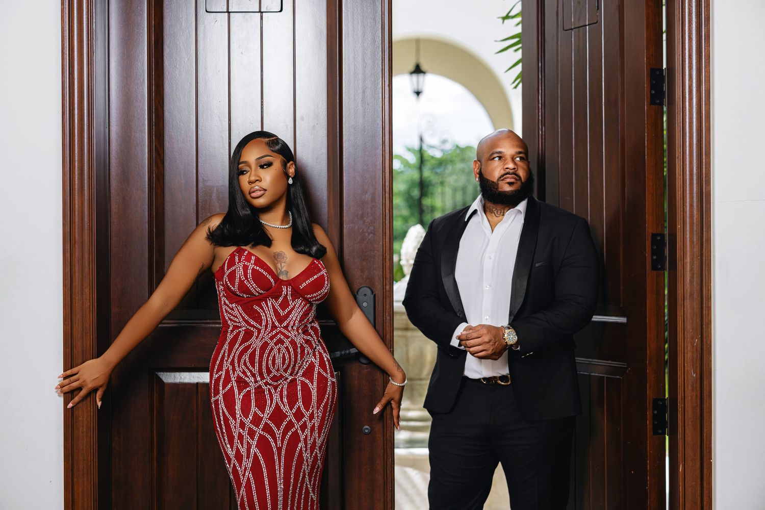 Stylish couple standing in ornate wooden doorway, woman wearing stunning red beaded evening gown.