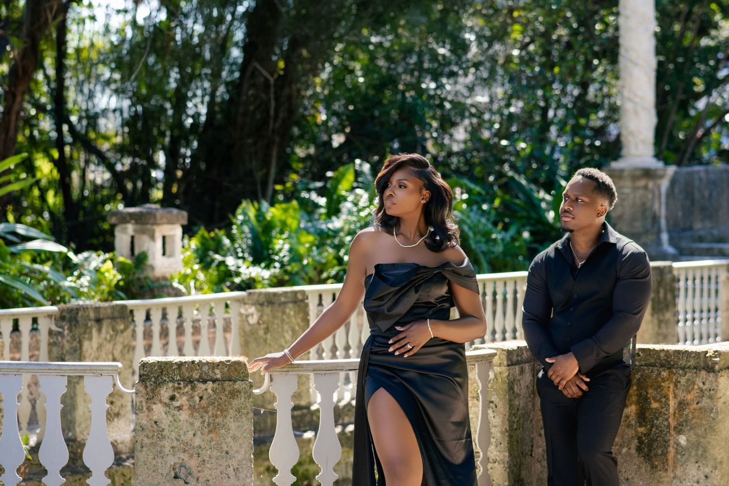 Woman in black slit gown posing with man on stone balustrade in lush garden.