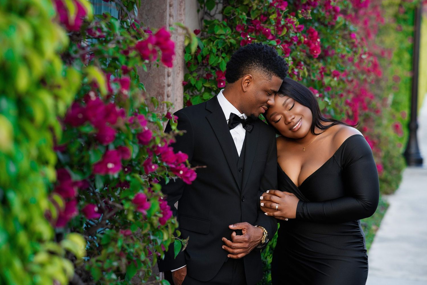 A romantic couple in black formal wear embrace tenderly against a colorful floral garden wall backdrop.