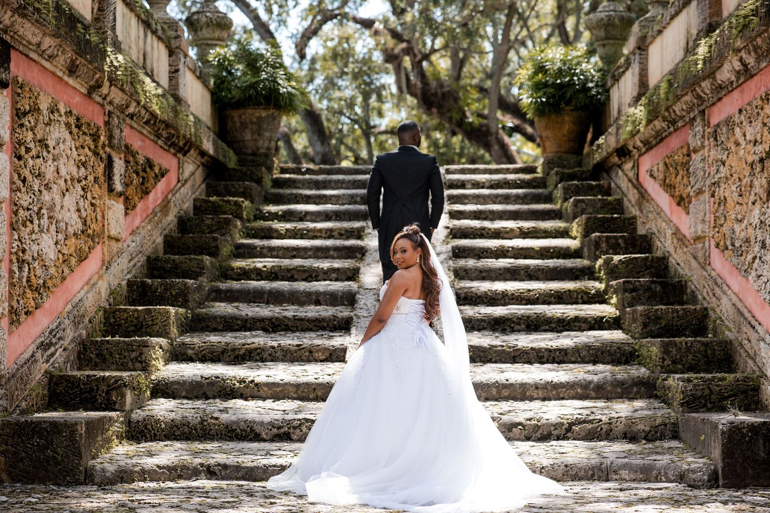 Bride in white gown sits on stone steps as groom walks up at historic venue.