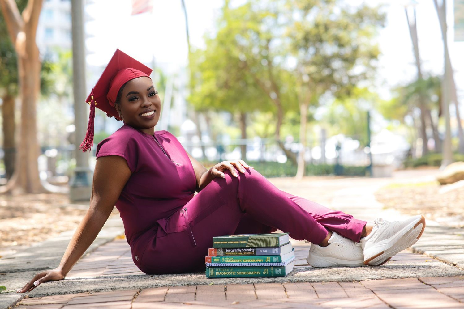 Smiling Black woman in magenta scrubs and graduation cap sits relaxed on brick path beside sonography textbooks outdoors.