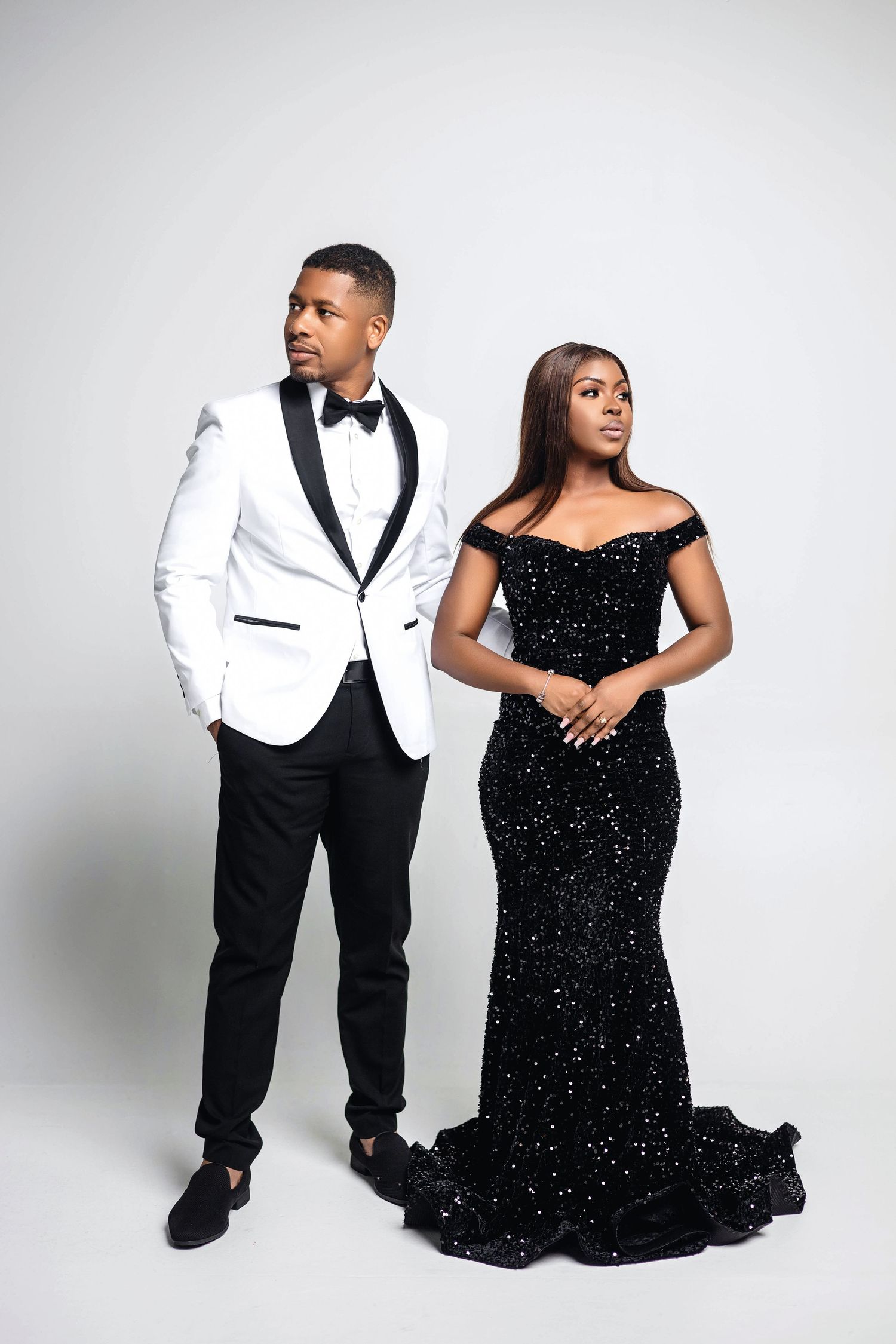 Couple in formal black and white attire posing on white background, woman in sequin gown, man in tuxedo.