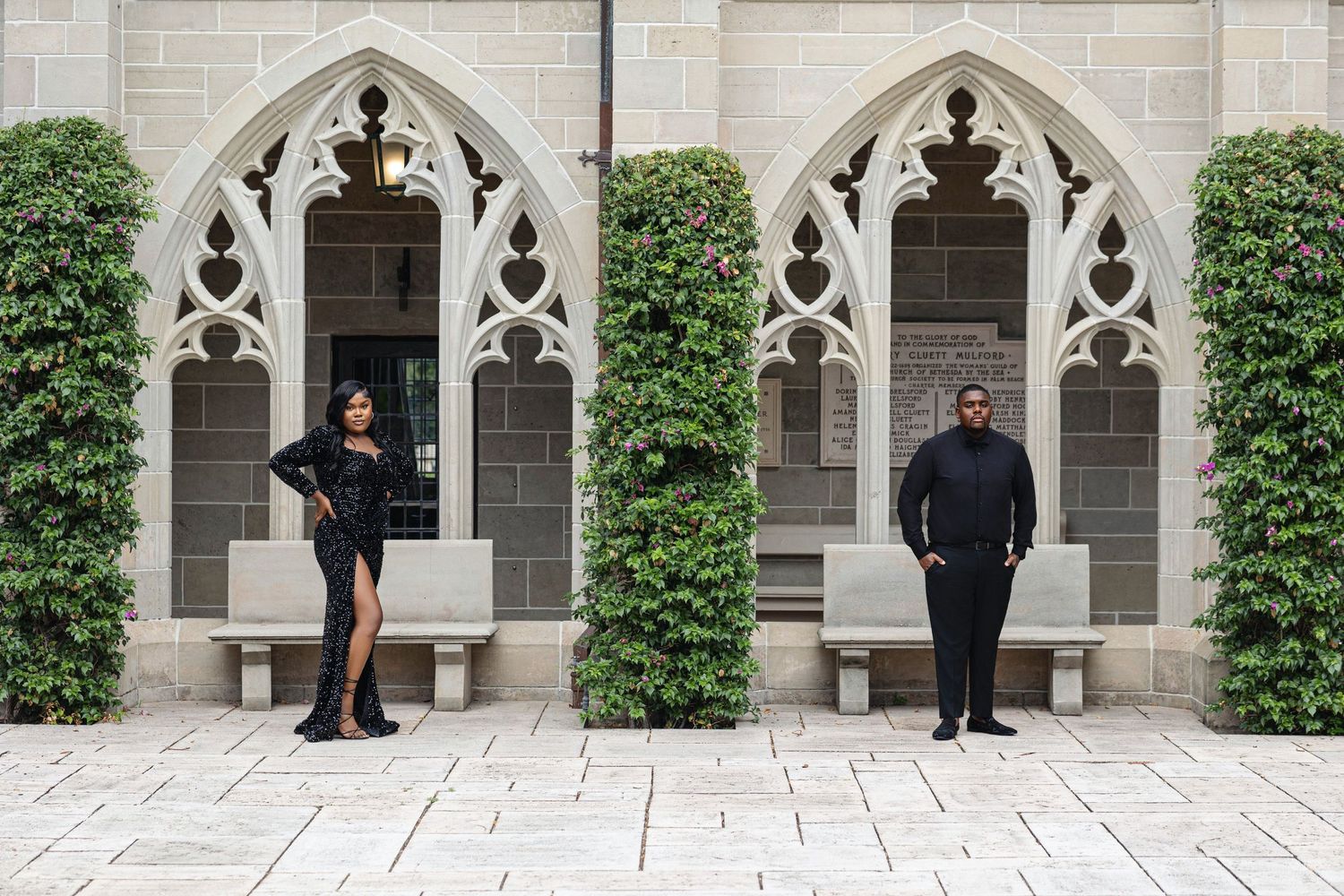 Elegant couple in matching black outfits posing before Gothic stone arches with lush ivy.