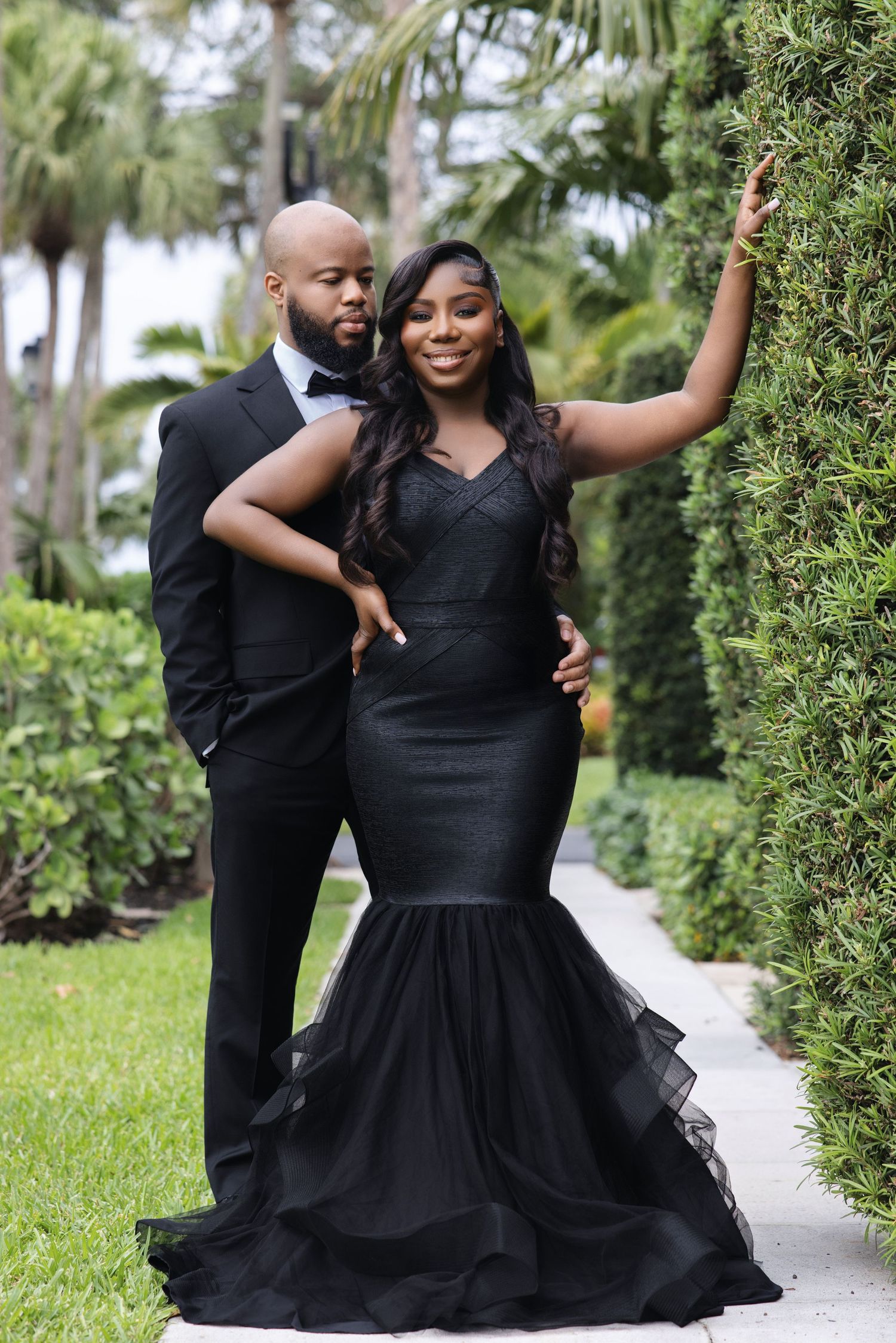 Black couple in matching black formal attire posing beside lush green hedges outdoors.