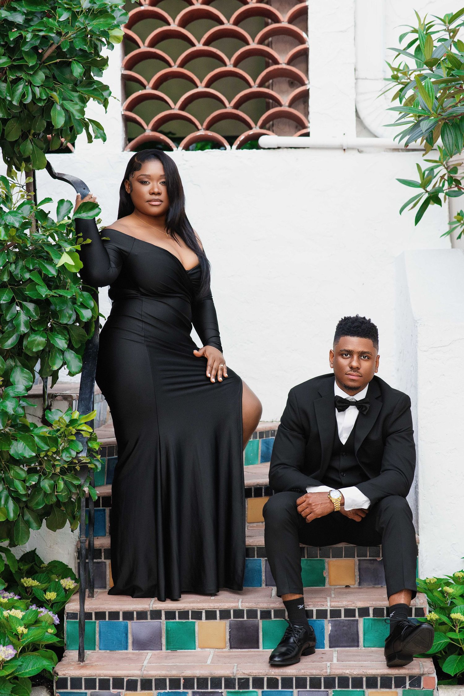 A glamorous couple in black formal wear pose dramatically on mosaic-tiled steps surrounded by lush greenery.