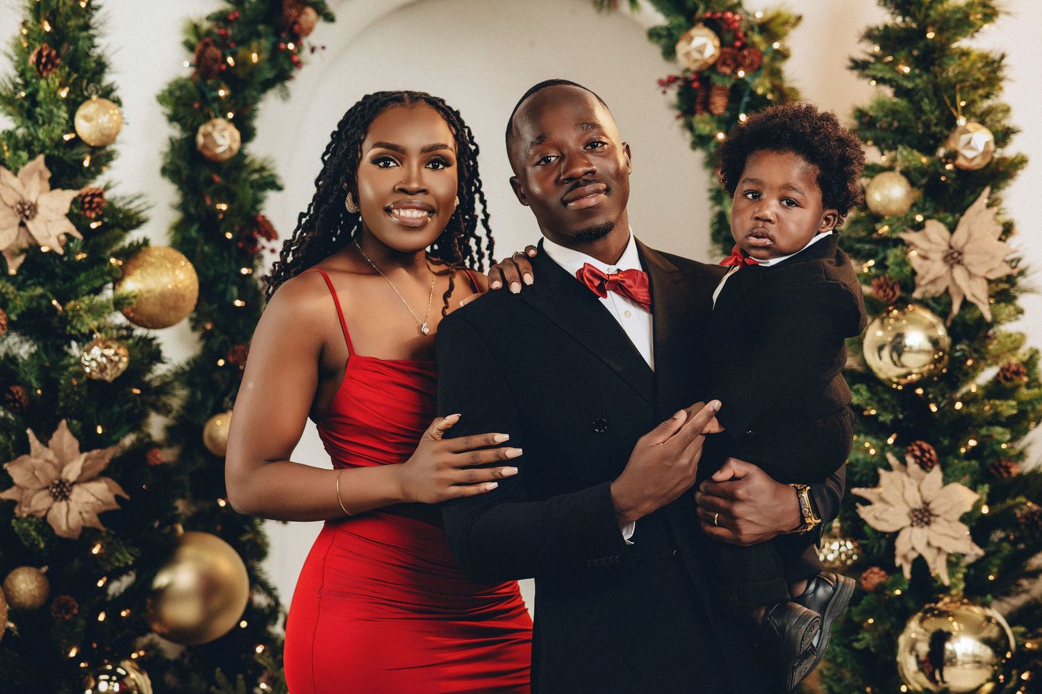 Family of three in matching red and black holiday outfits posing before Christmas trees.