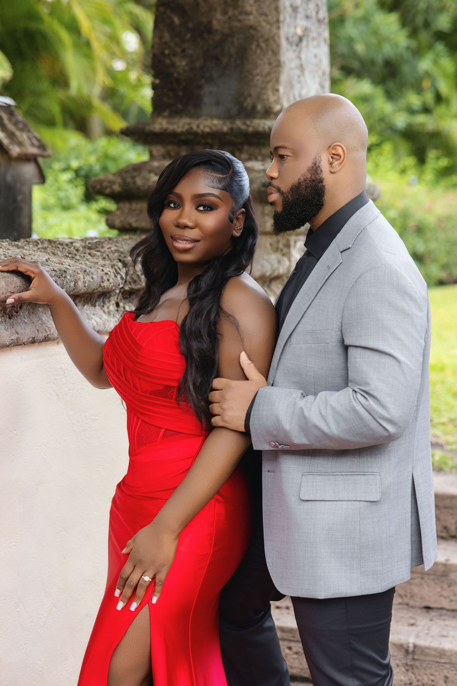 Woman in red strapless gown and bald man in gray blazer posing by stone wall outdoors.