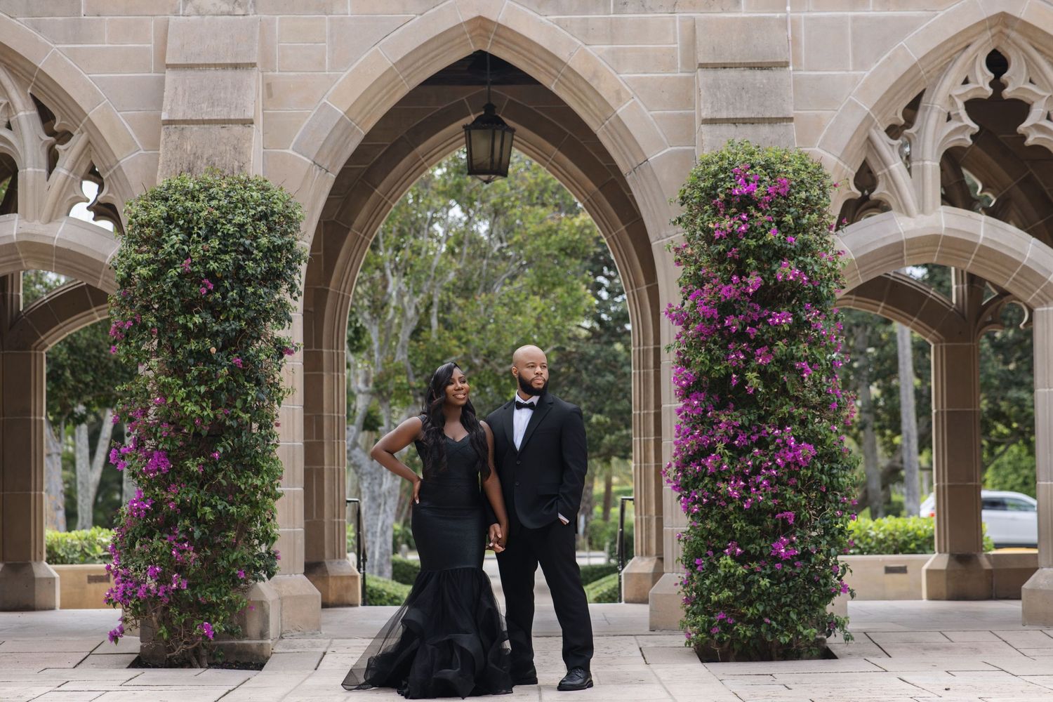 Elegant couple in black formal wear posing under gothic stone arches with purple bougainvillea flowers.