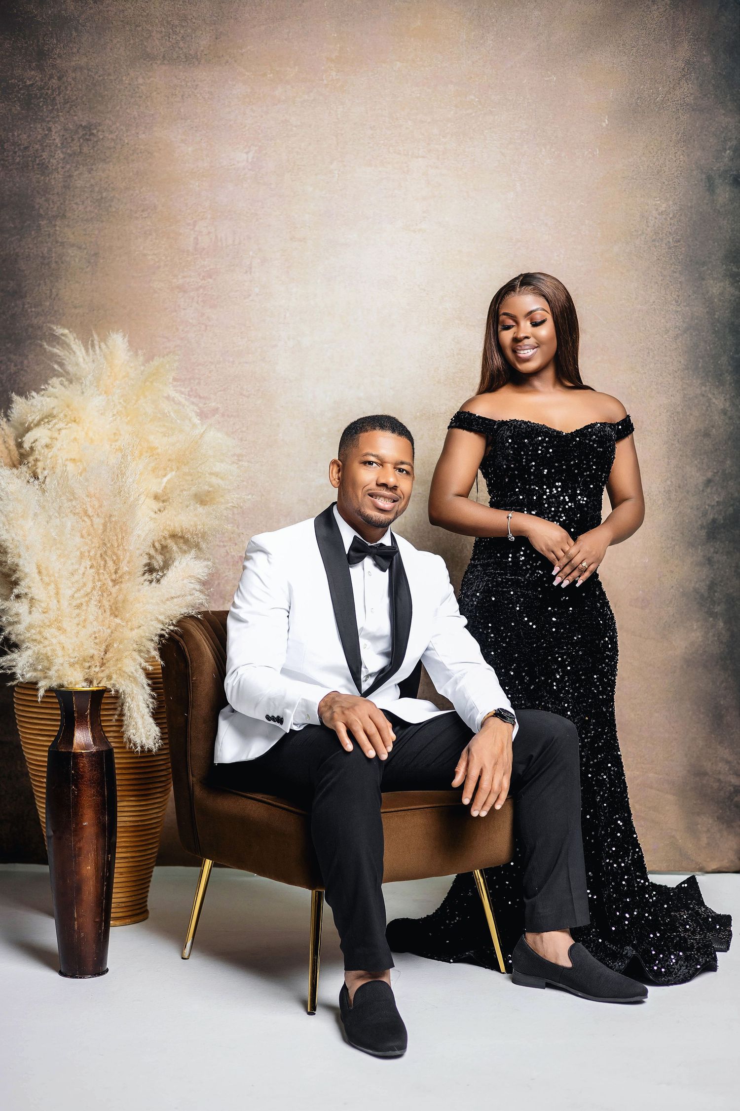 Elegant couple in studio, man in white tuxedo seated, woman standing in black sequin gown.