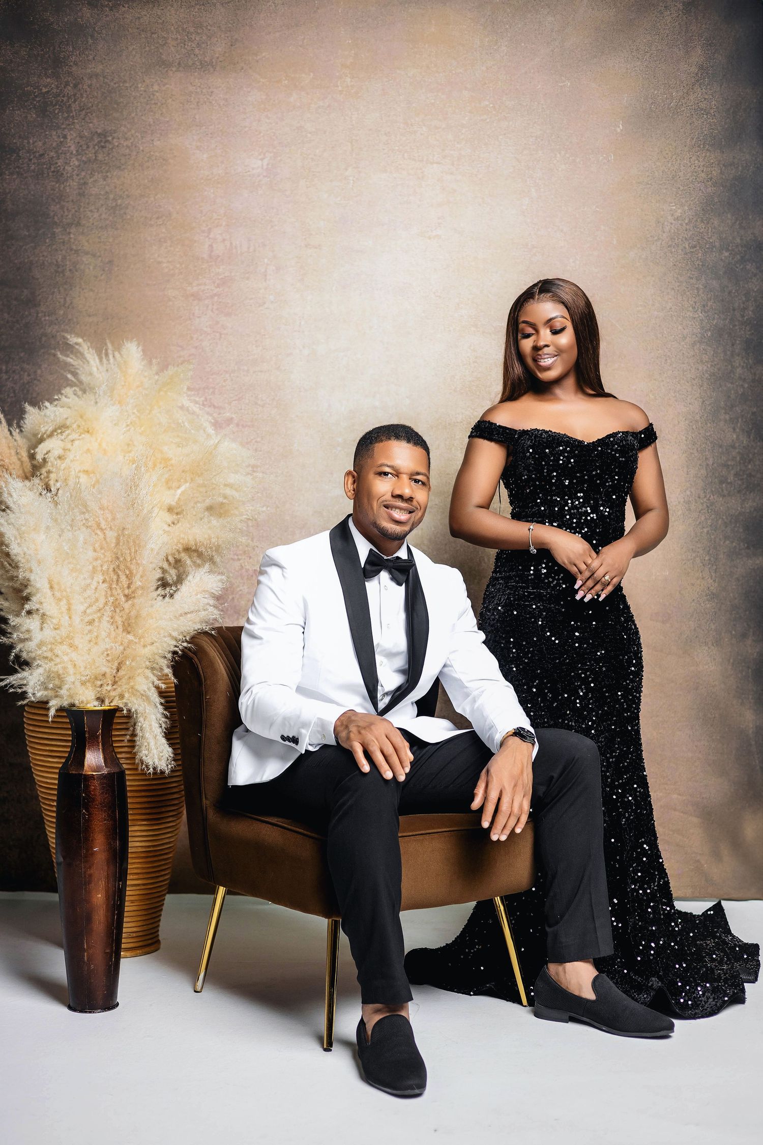 Elegant couple in formal wear, man seated in velvet chair wearing white tuxedo, woman in black sequin gown.