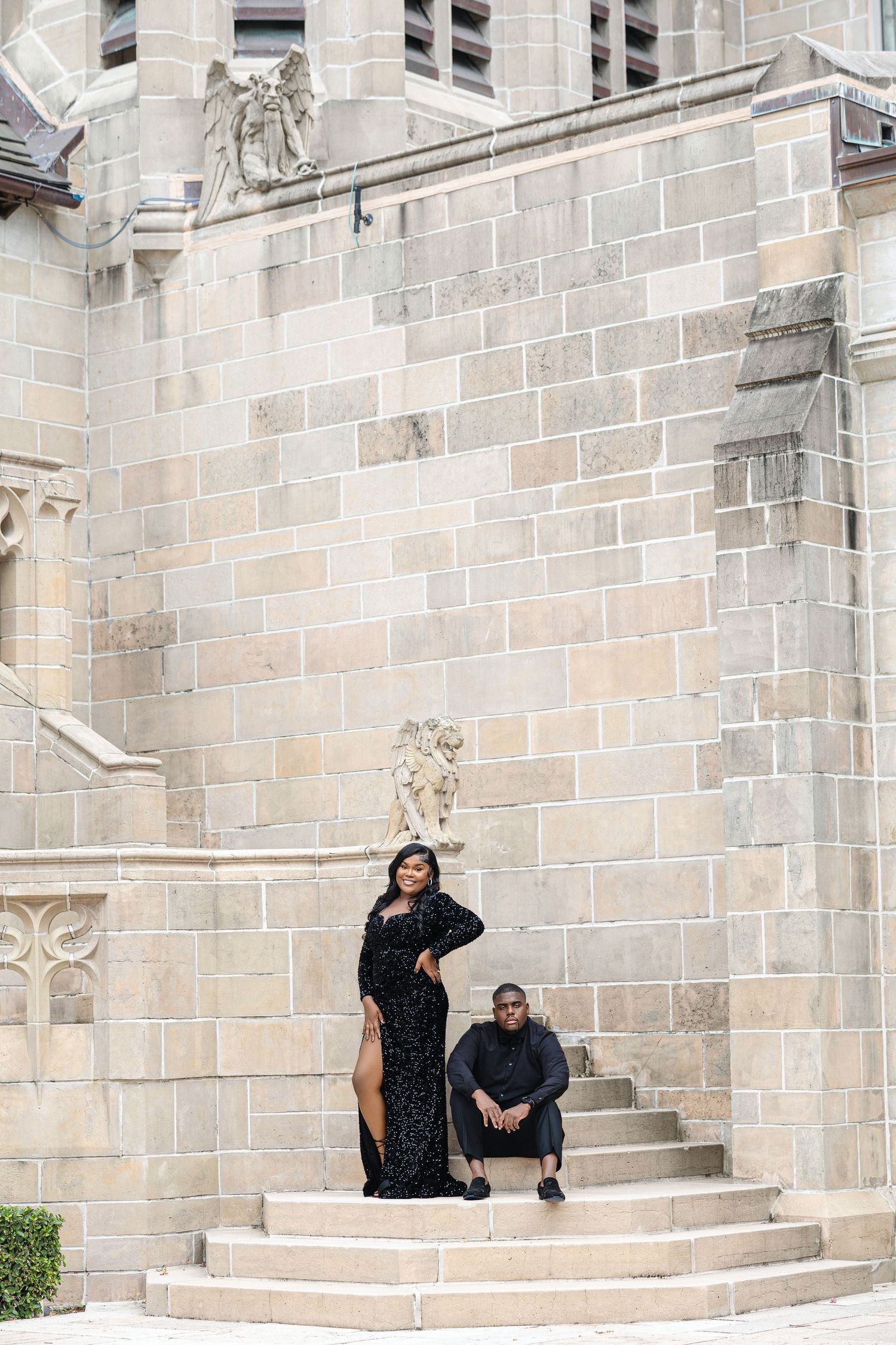 Couple in black formal wear posing on stone steps of a grand historic building.