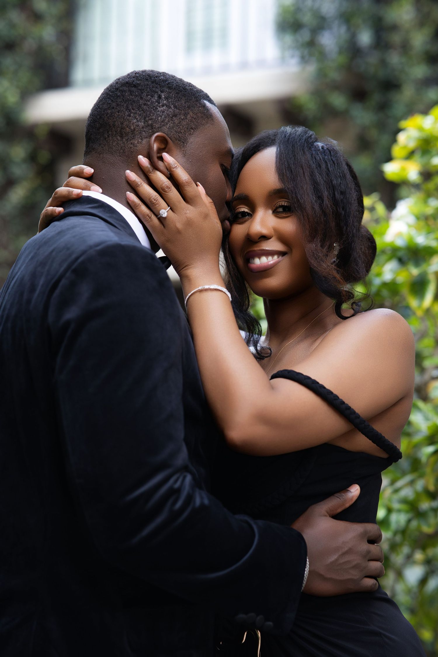 A couple in elegant black attire embrace outdoors, the woman smiling warmly while holding her partner's face tenderly.
