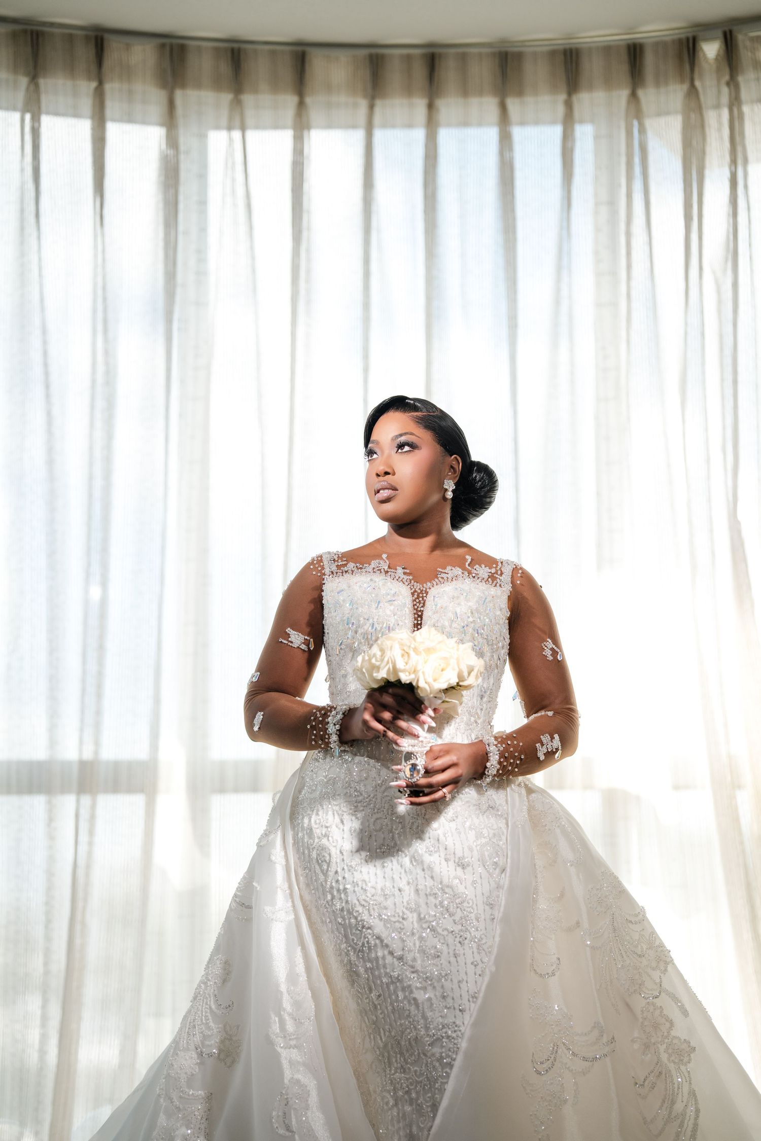 Elegant bride in lace long-sleeve wedding gown holding white bouquet, standing before sheer curtains in soft natural light.