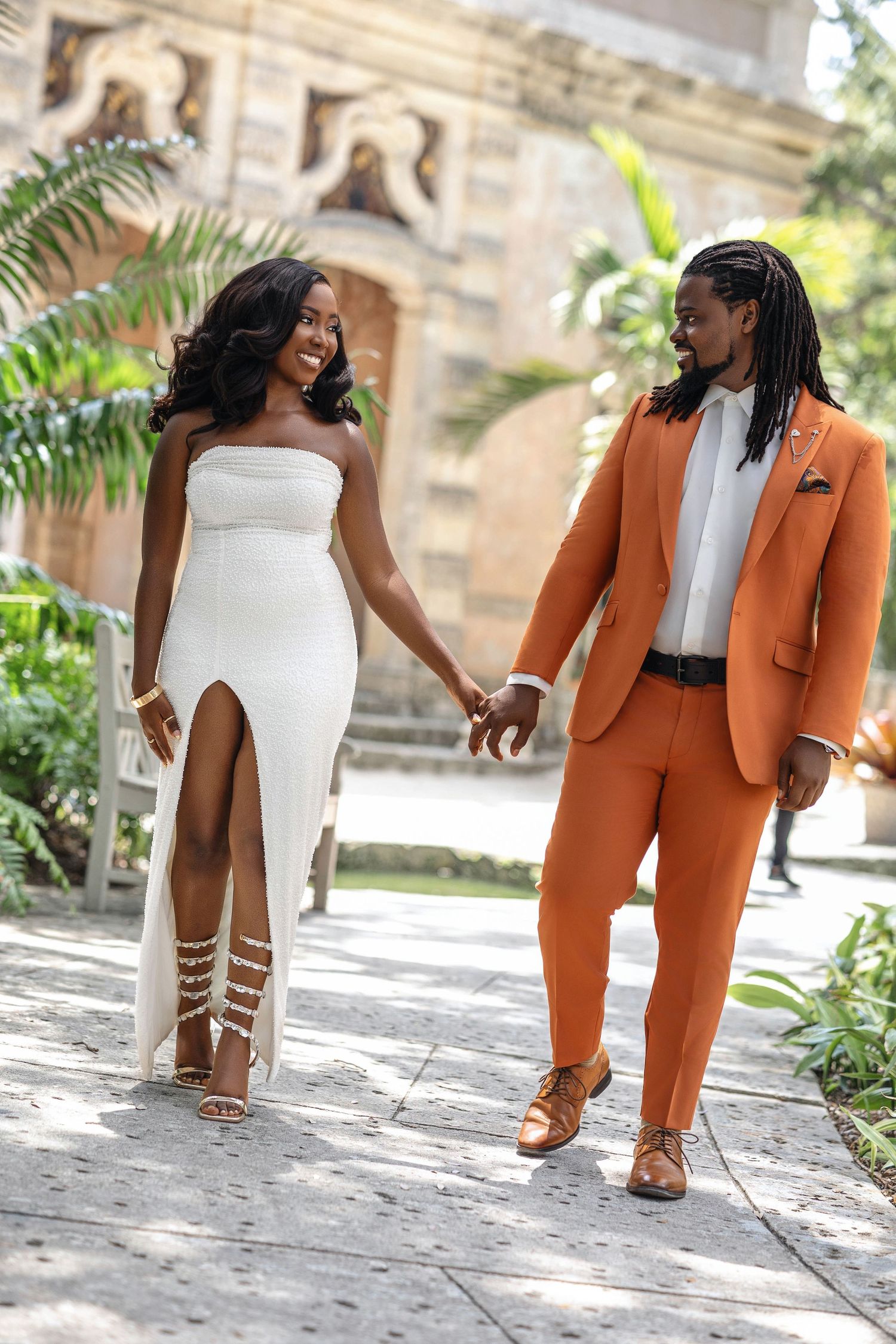 Elegant couple holding hands walking through garden, woman in white gown and man in orange suit.