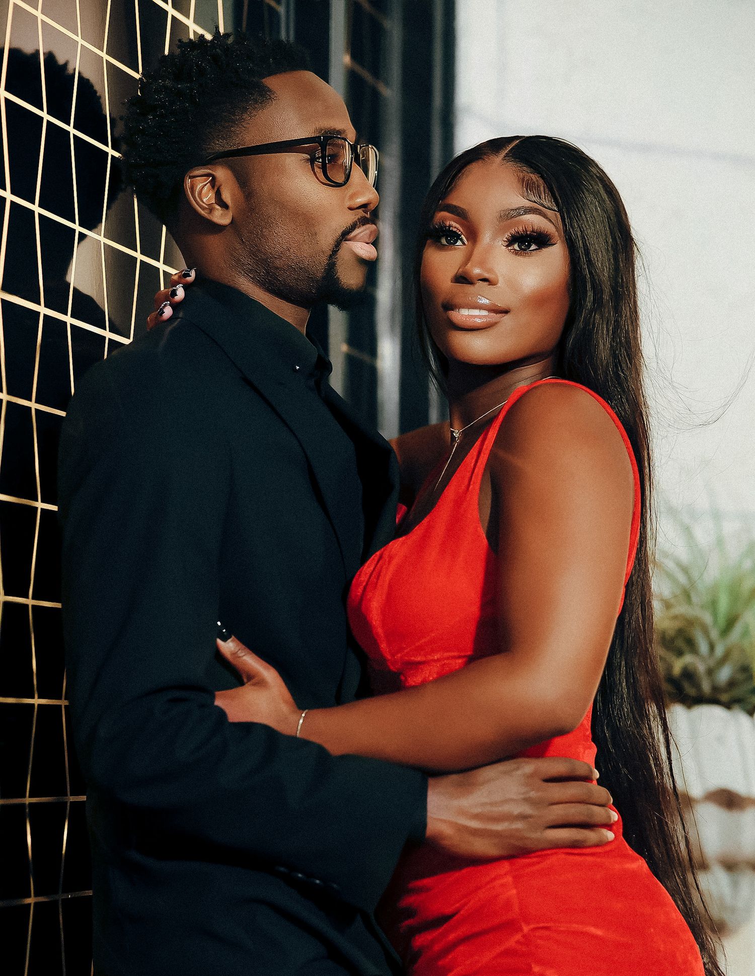 Close-up of stylish couple in red dress and black suit posing against a geometric gold and black wall.