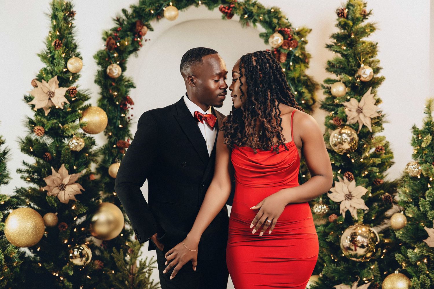 Couple in elegant red dress and black tuxedo gazing at each other by Christmas trees.