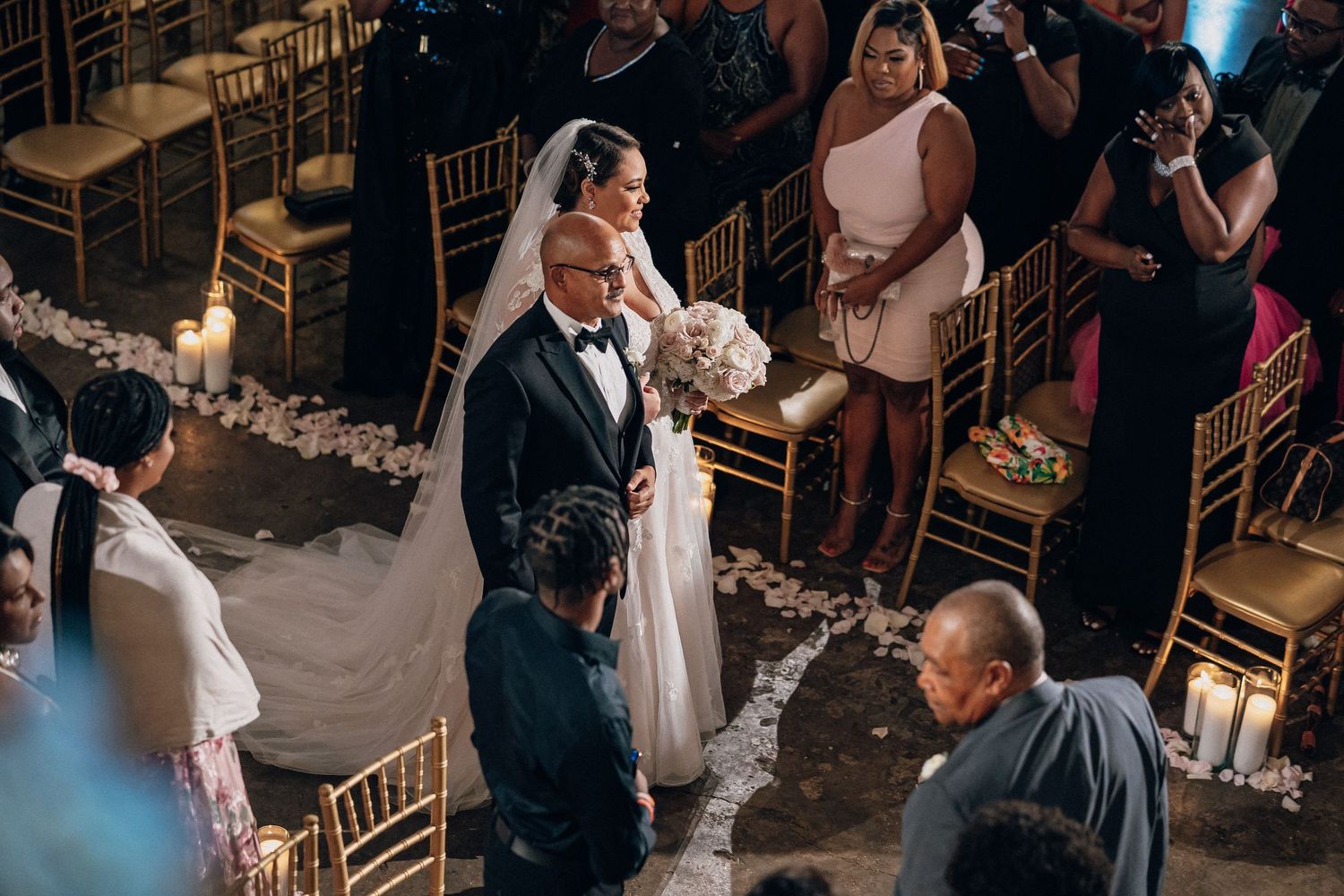 Bride in white gown walks down aisle with escort at elegant candlelit wedding ceremony with gold chairs.