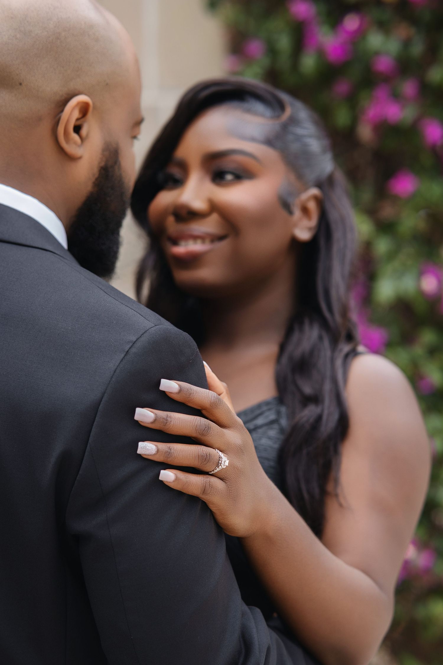 Smiling woman in black dress with diamond engagement ring resting hand on partners suited shoulder.