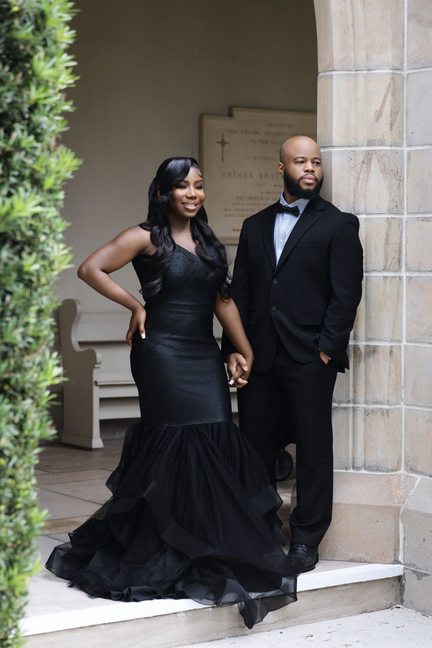 Elegant Black couple in black formal wear posing at a stone archway with a cross.