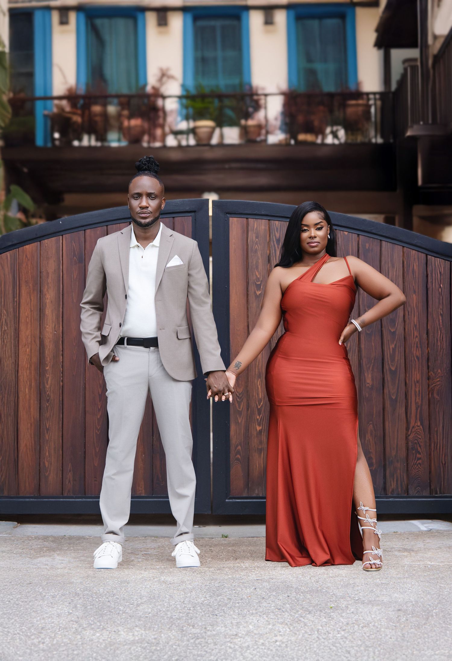 A Black couple holds hands posing confidently before wooden gates, dressed in elegant formal attire.