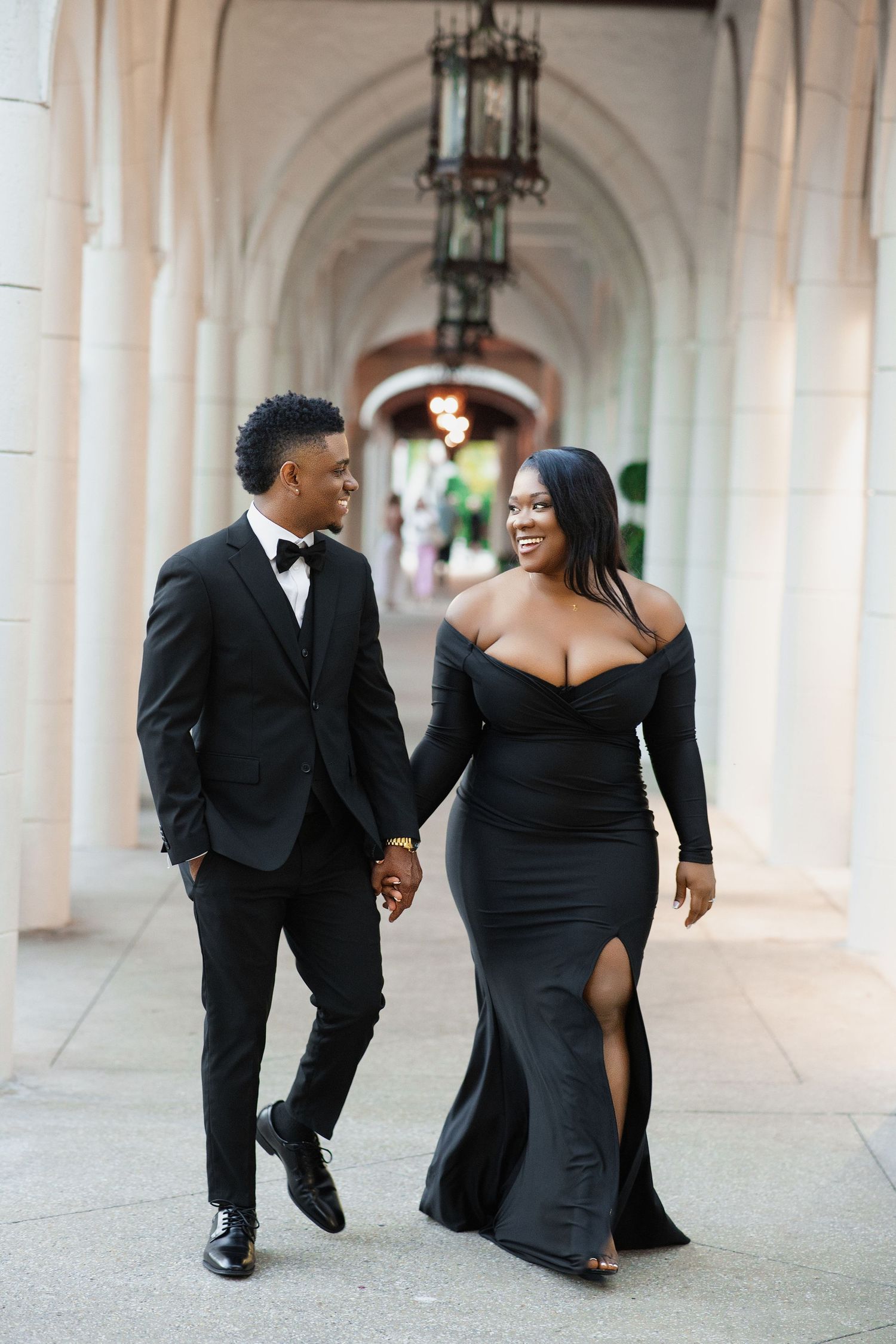 Joyful Black couple in elegant black formal wear walking hand-in-hand under arched columns.