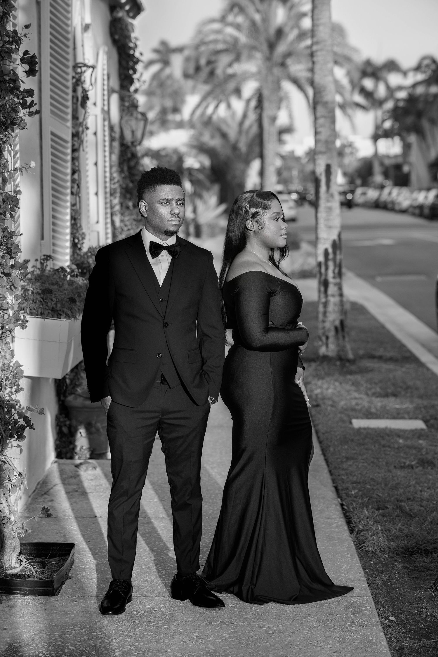 A stylish couple in black formal attire pose on a palm-lined sidewalk in this classic black and white portrait.