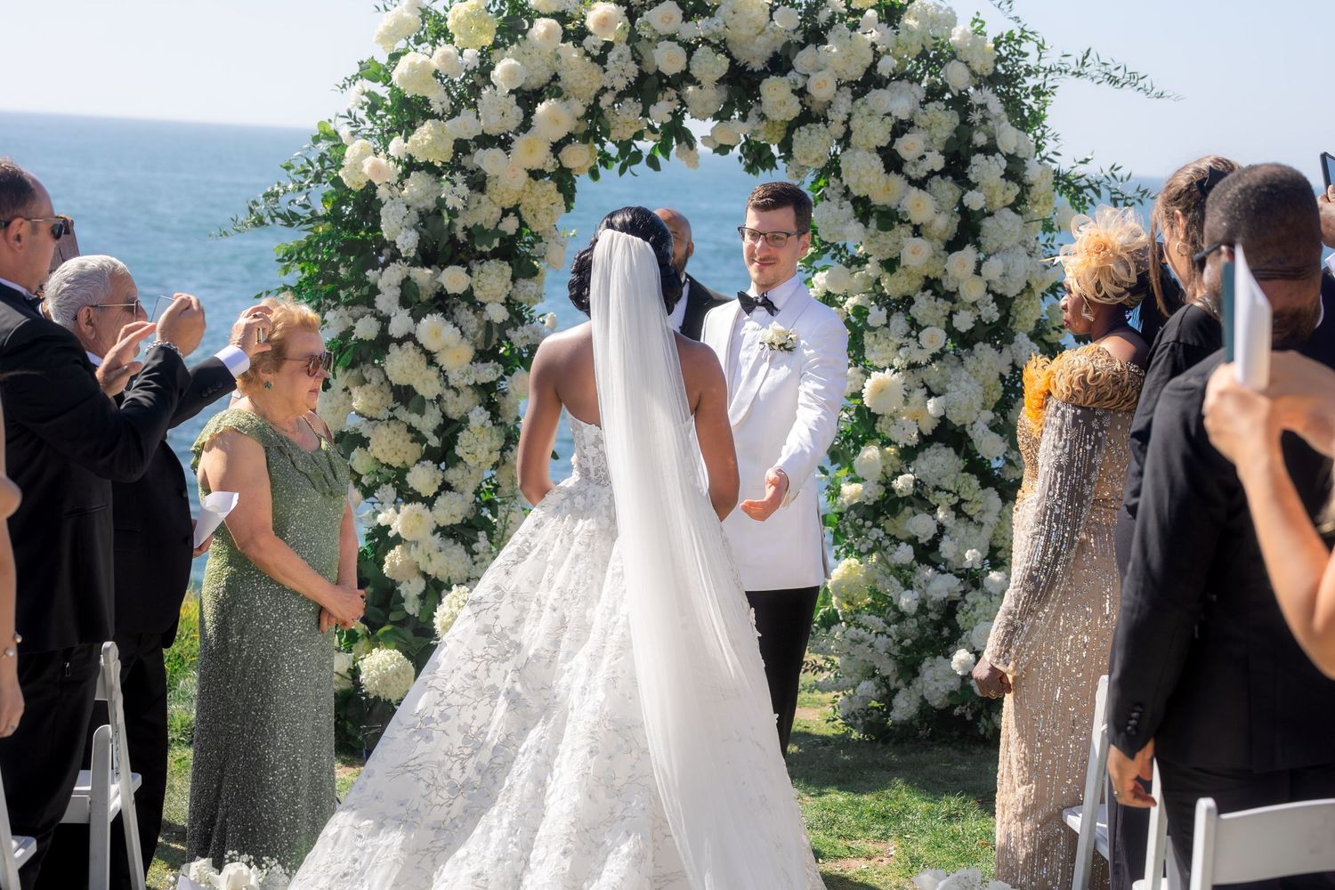 Outdoor wedding ceremony with bride and groom at floral arch overlooking ocean, surrounded by guests.