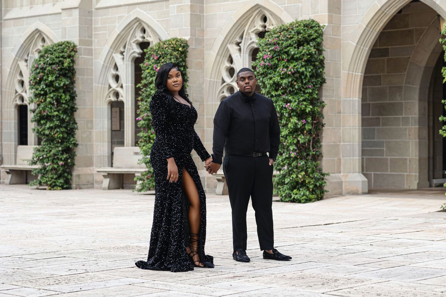 Engaged couple in coordinating black outfits holding hands in Gothic stone courtyard with arched walkways.
