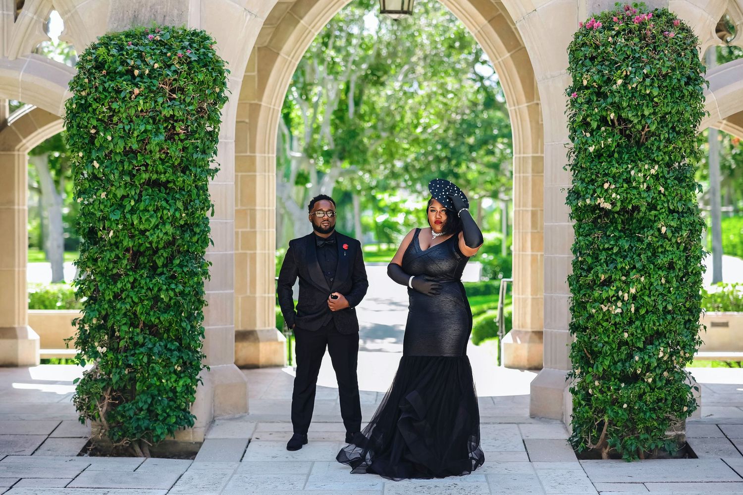 A couple in black formal wear stand apart beneath lush ivy-covered arches in a manicured garden.