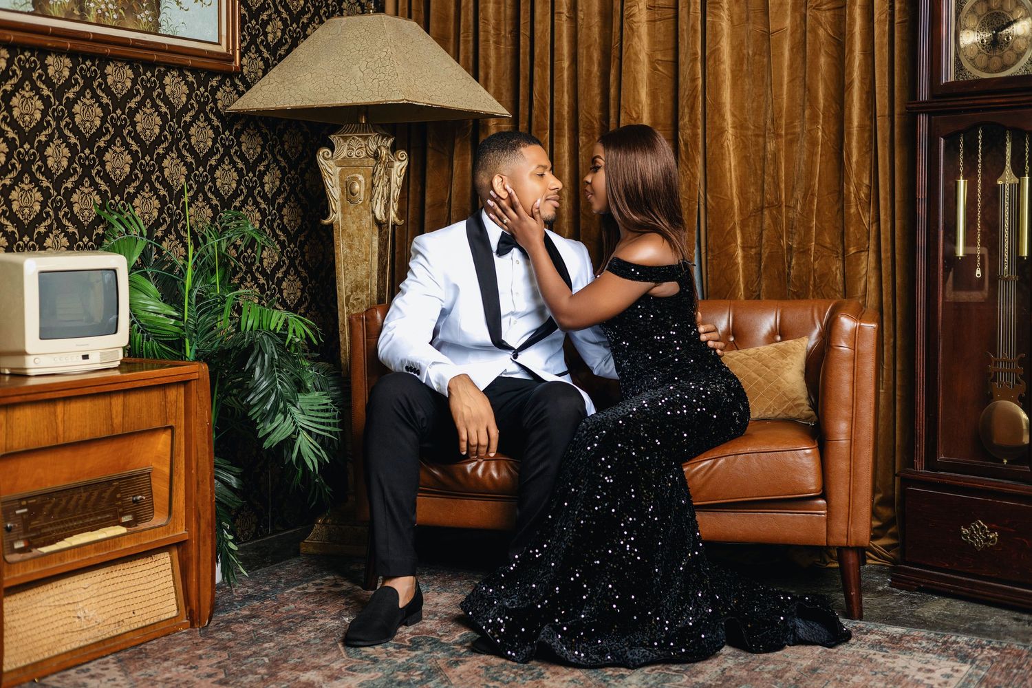 Couple in elegant formalwear sharing intimate moment on brown leather sofa in vintage-decorated room.
