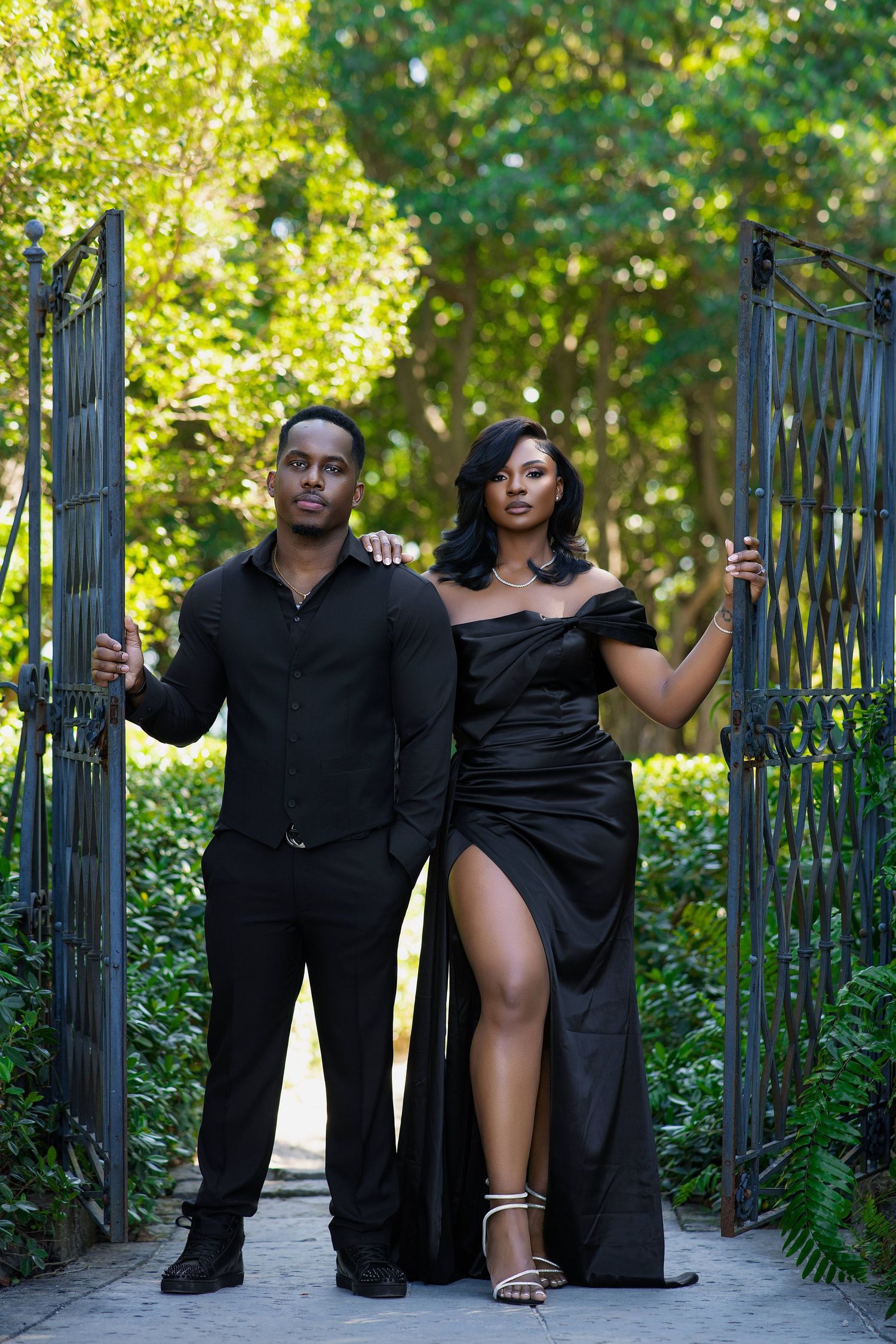 Black couple posing confidently at ornate iron garden gate, woman in black off-shoulder gown with slit.