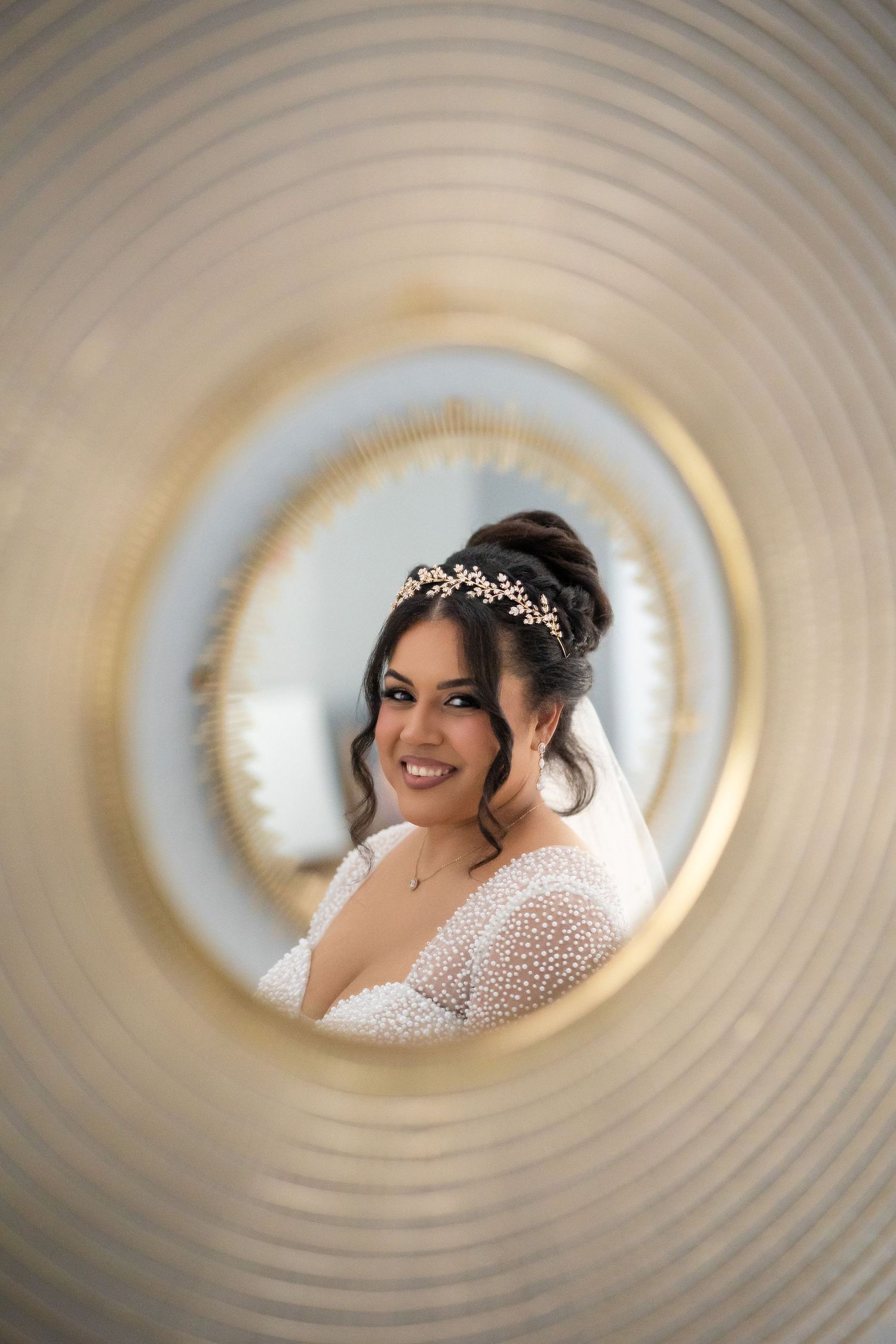 Bride in pearl wedding dress with floral headpiece smiling, reflected in ornate gold circular mirror.