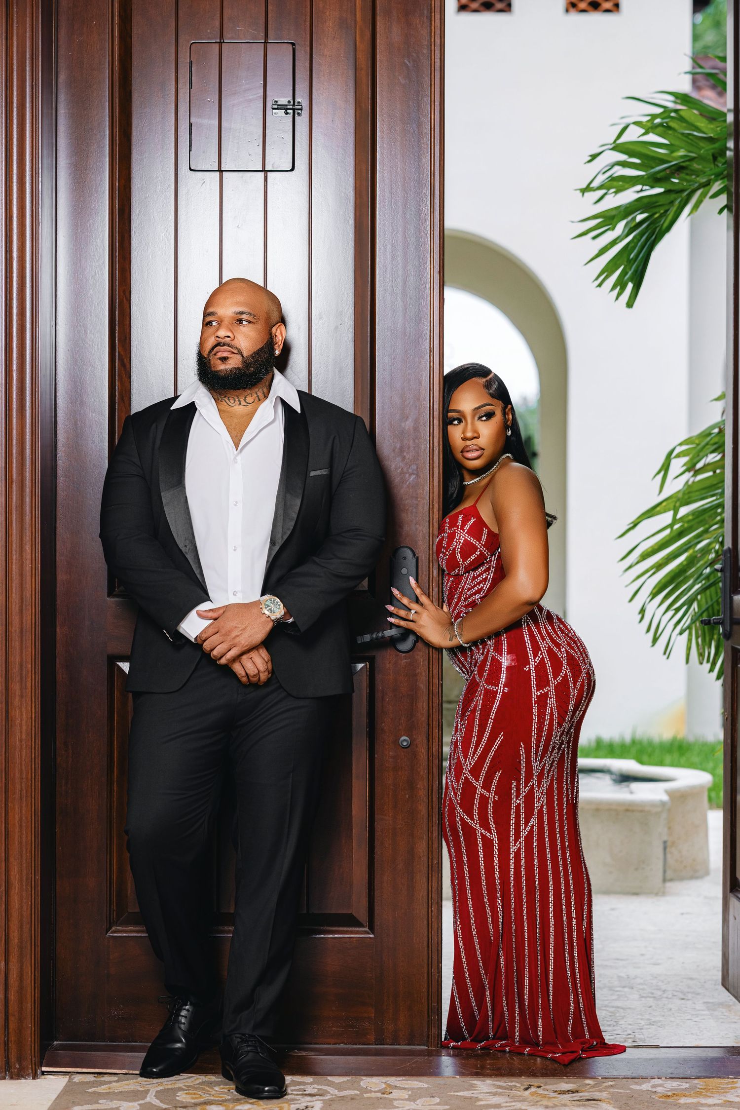 Couple poses at grand wooden entrance doors, man in black tuxedo and woman in red beaded evening gown.