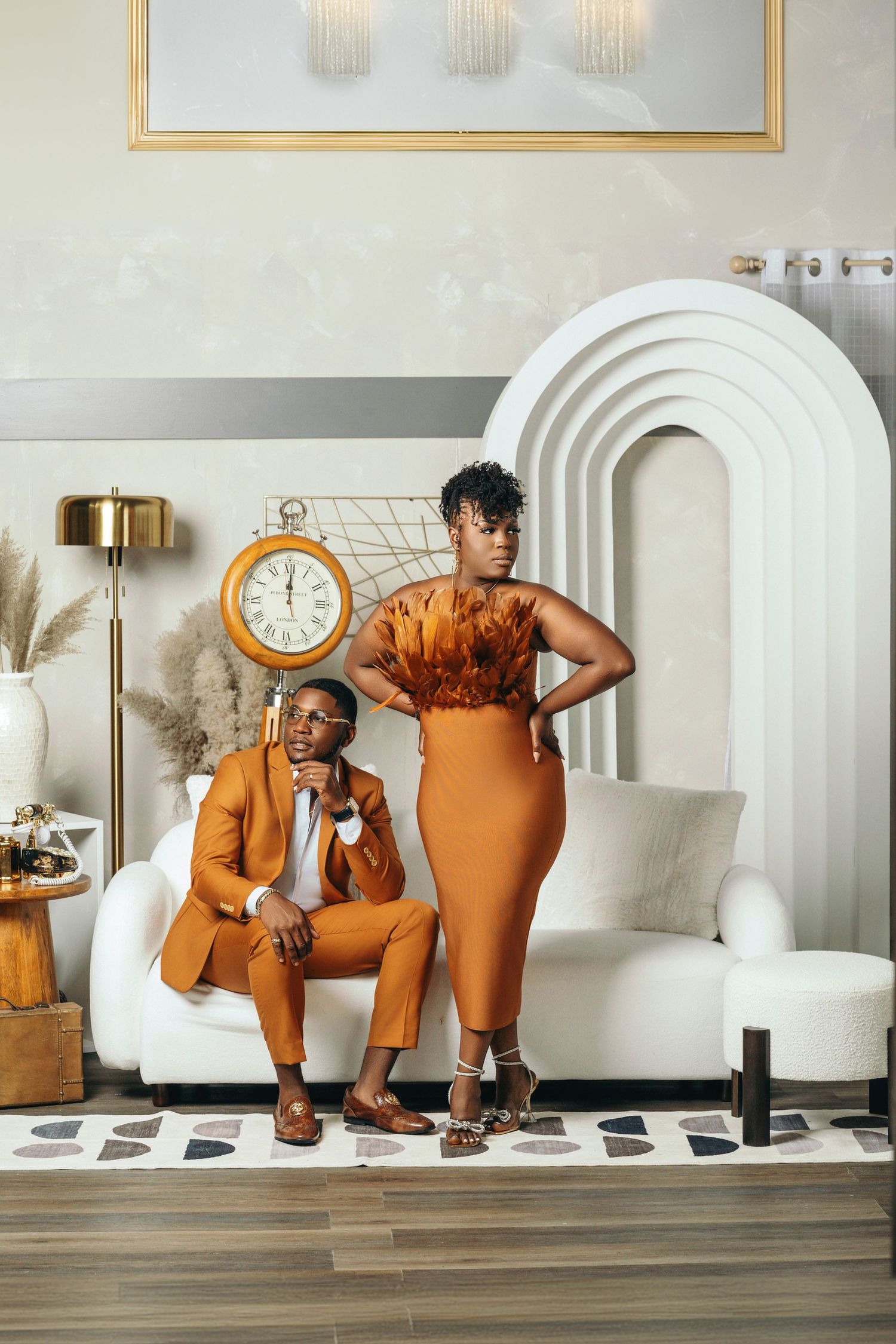 Couple in matching burnt orange outfits posing elegantly in a modern white interior with arch decor and gold clock.