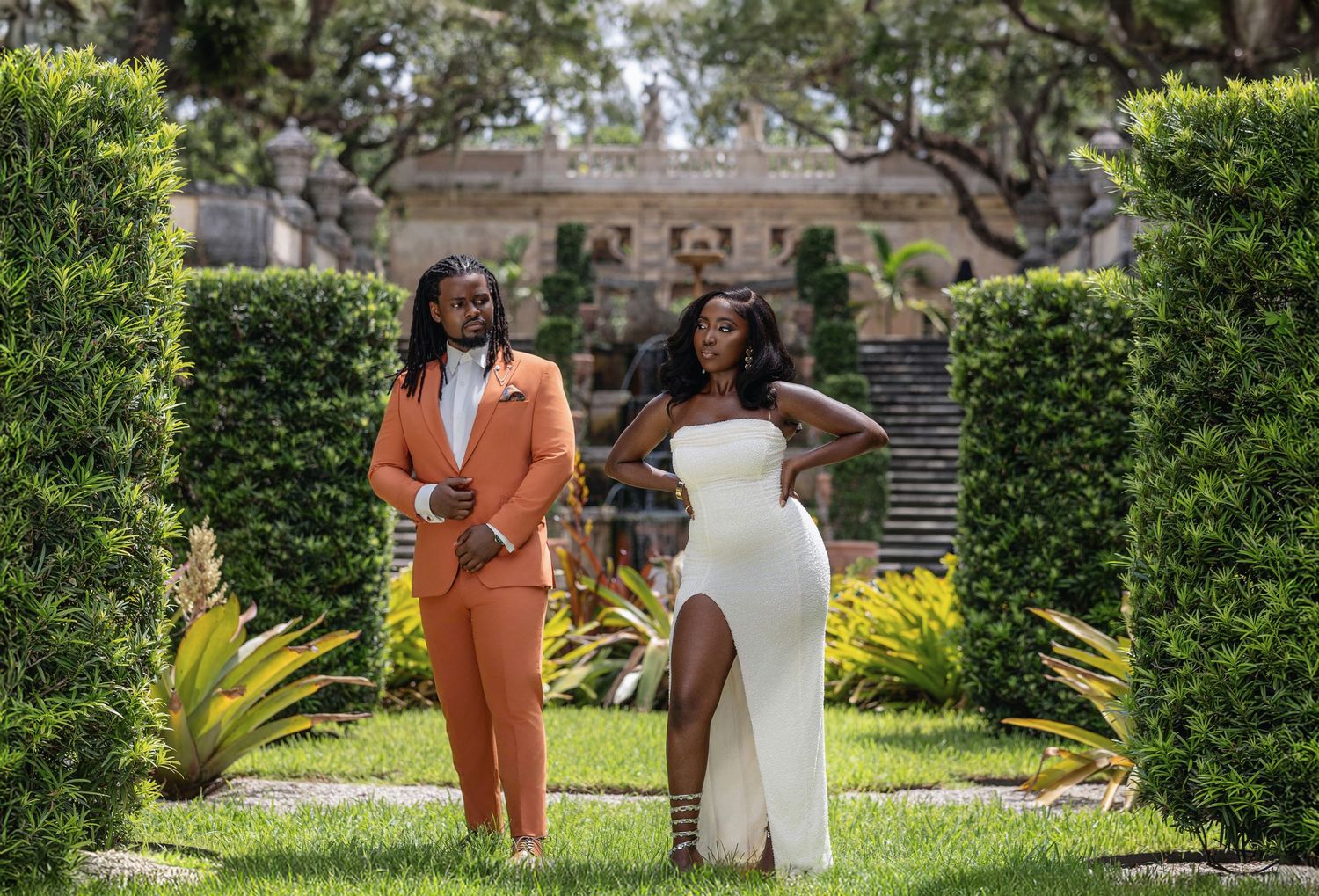 Stylish engaged couple posing between manicured hedges at historic estate in coordinated outfits.