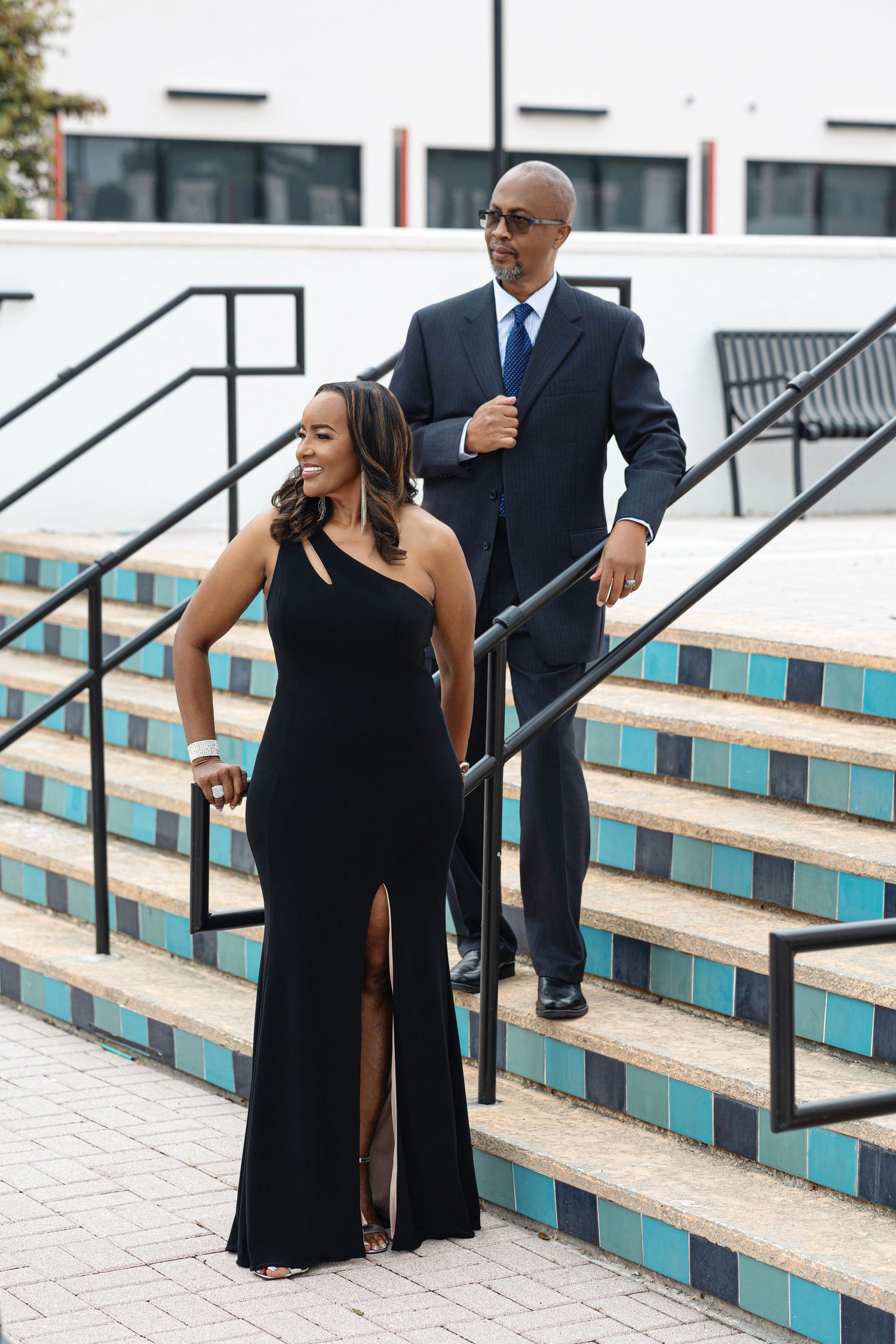 Couple in elegant black formal attire walking down decorative tiled stairs at outdoor location.