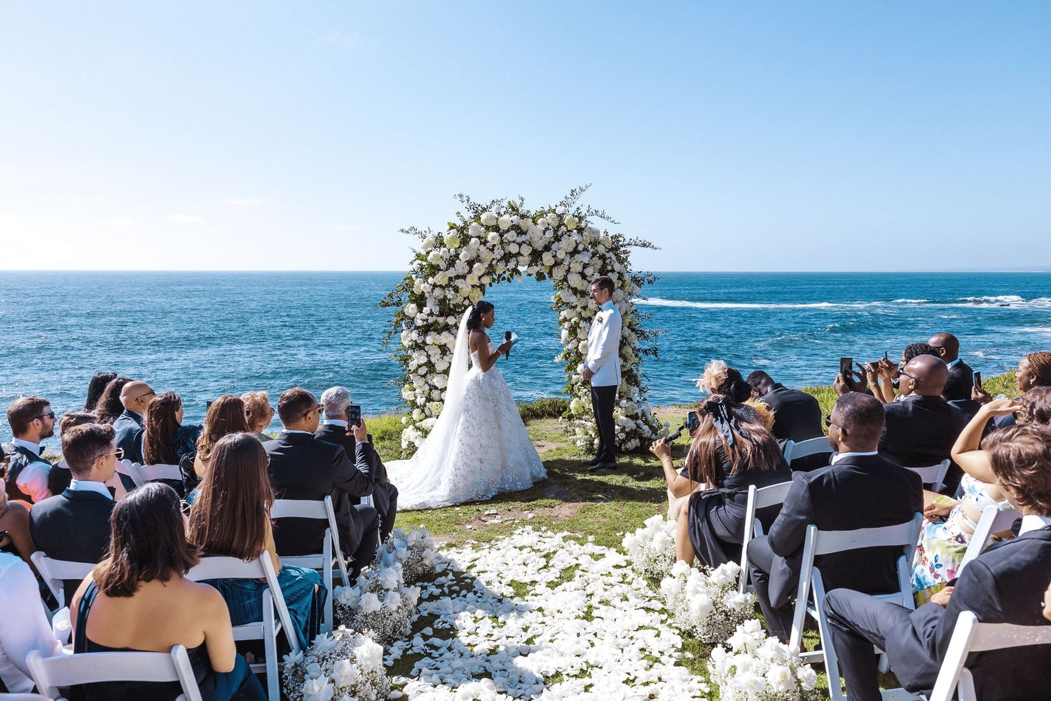 Outdoor oceanfront wedding ceremony with floral arch, bride and groom at altar, white chairs, and blue sea backdrop.