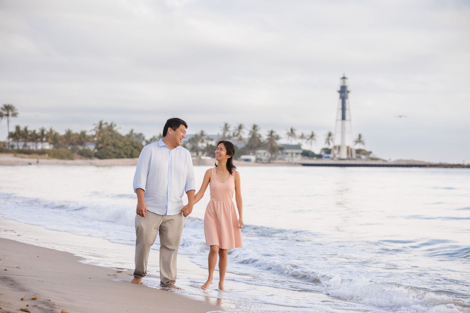 Couple holding hands walking on a tropical beach with a lighthouse in the background.