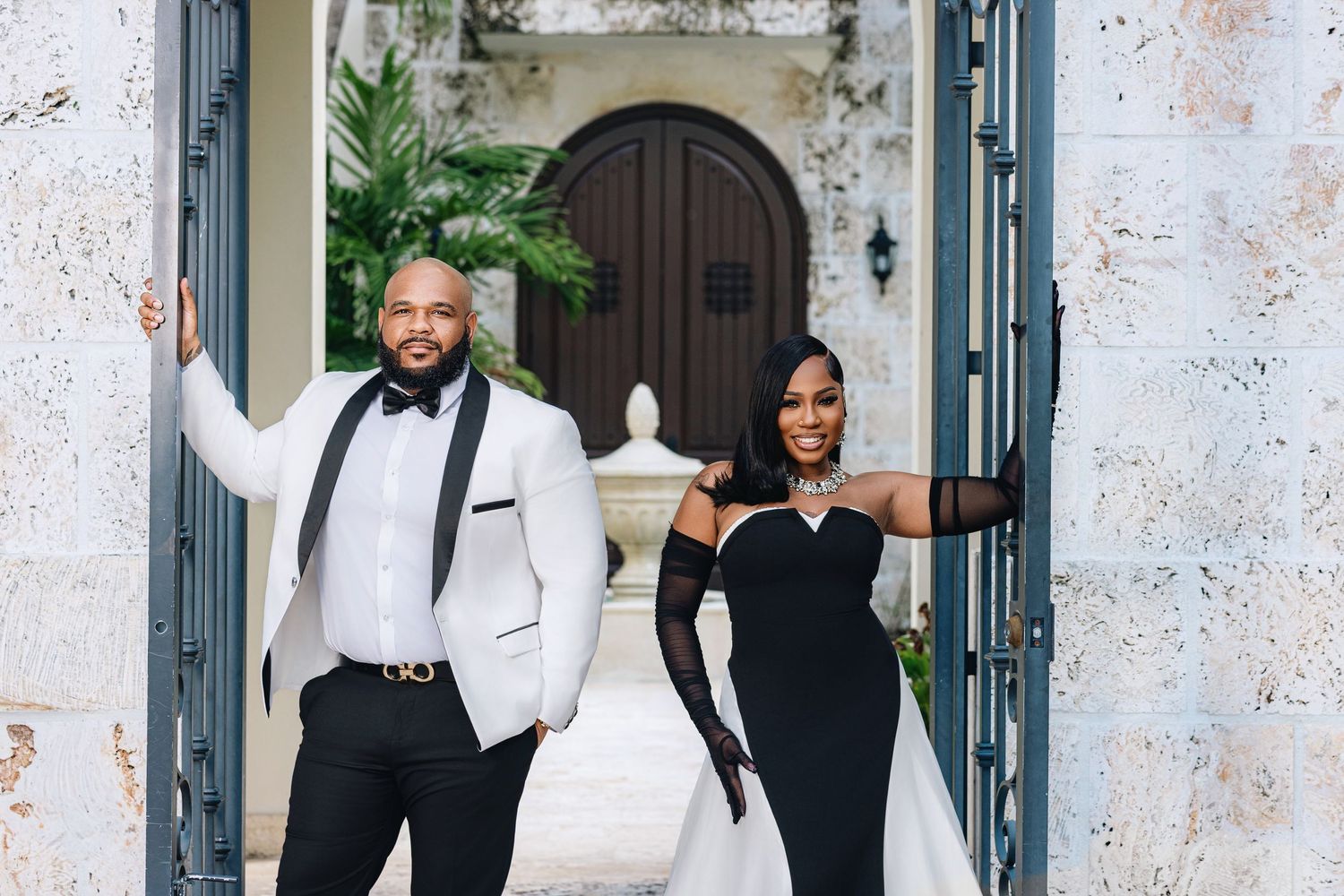 Smiling wedding couple standing in open decorative gates, groom in white tuxedo jacket, bride in black fitted gown.