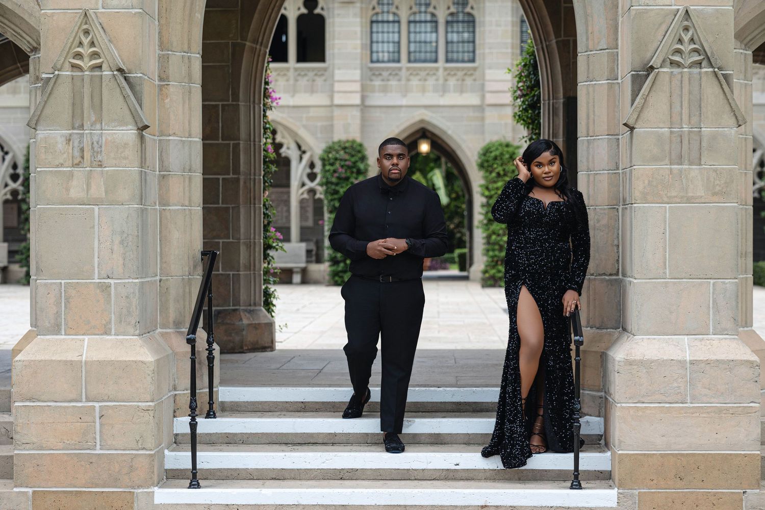 Couple in elegant black formal wear descending stone steps of a Gothic cathedral colonnade together.