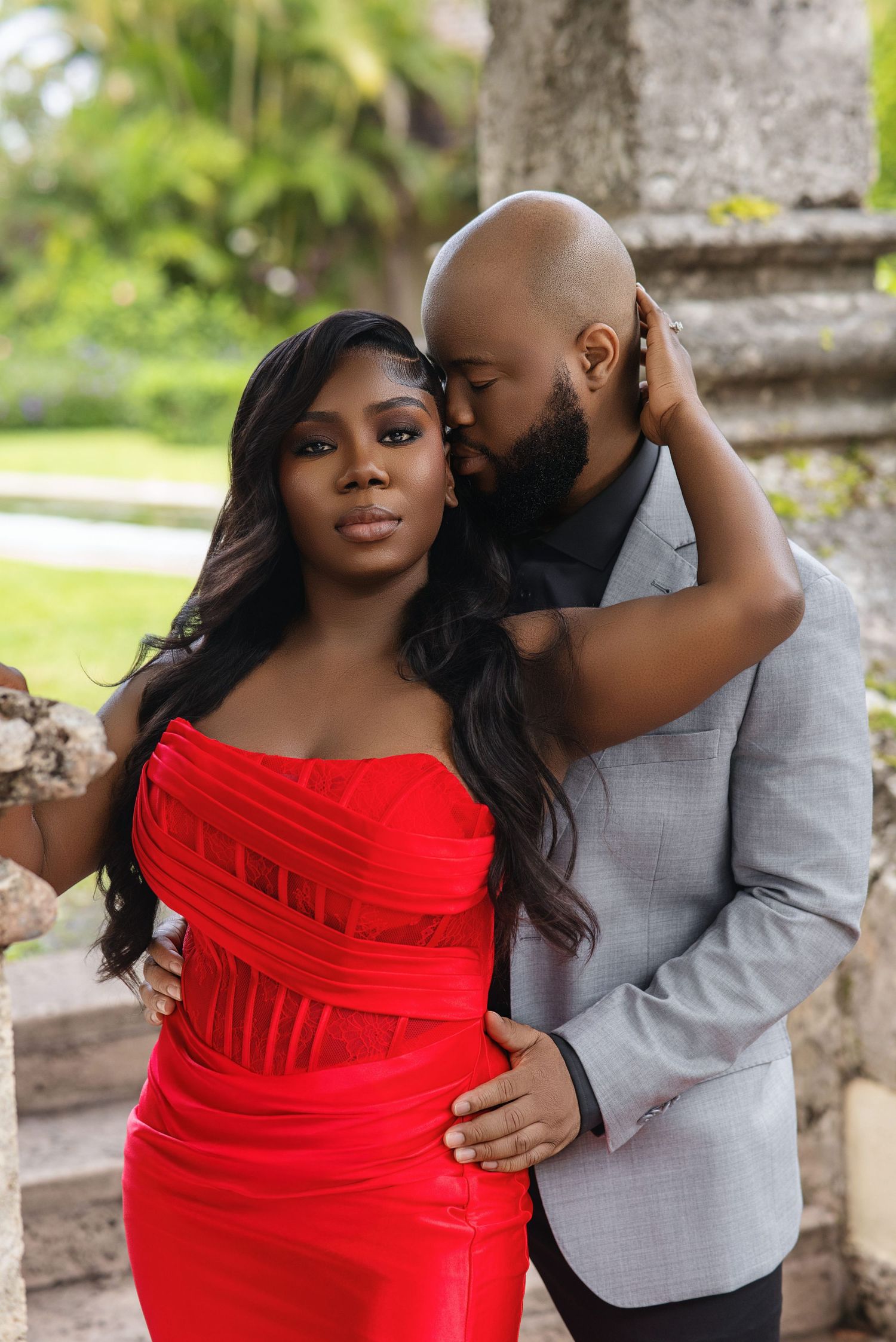 Romantic couple embrace outdoors, woman in red dress and man in gray suit kissing her cheek.